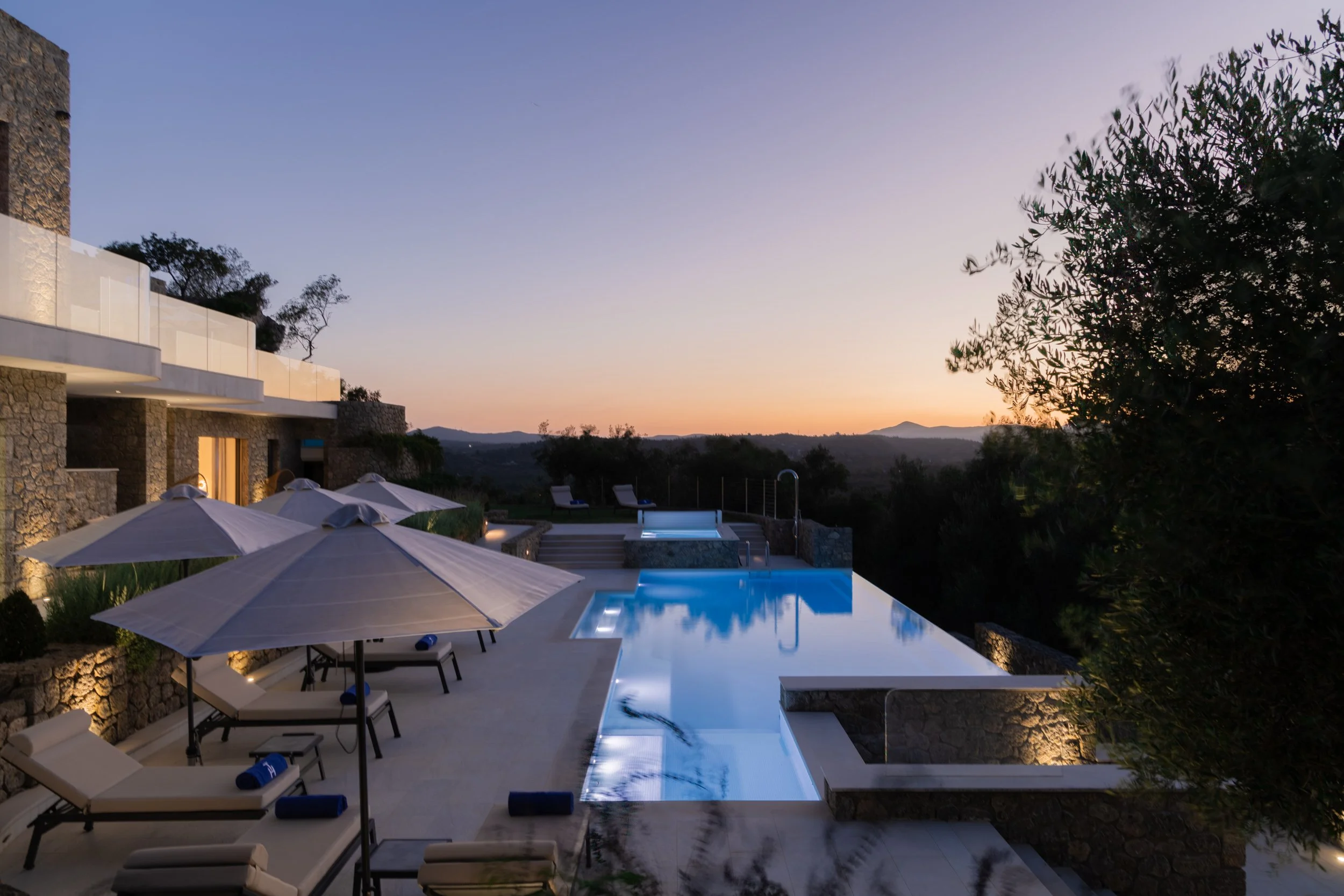 Luxury outdoor swimming pool at sunset with lounge chairs, umbrellas, and scenic mountainous landscape in the background.