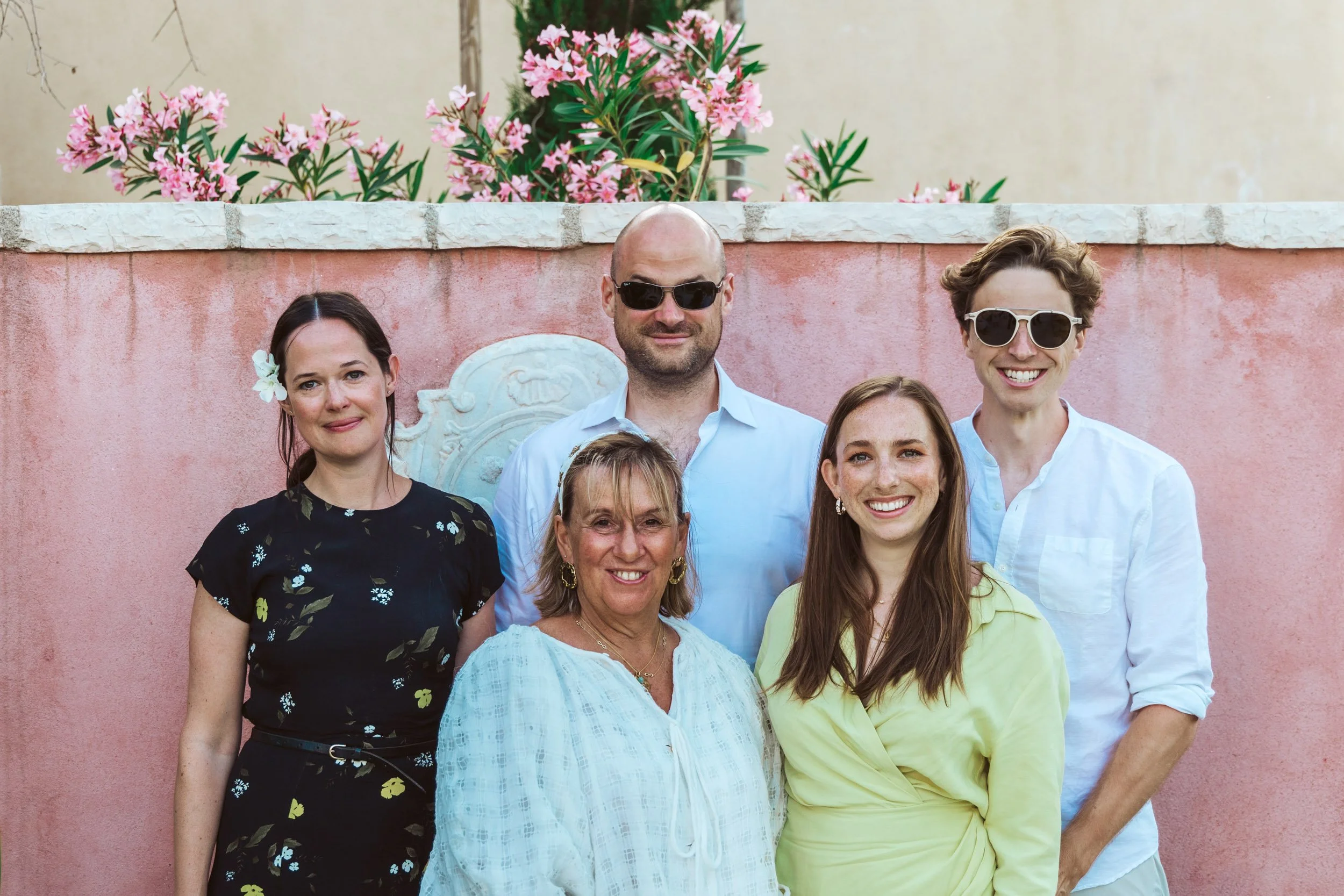 Group of six people smiling and posing together against a pink wall with flowering plants on top. They are dressed in casual and semi-formal clothing, some wearing sunglasses, outdoors during daytime.