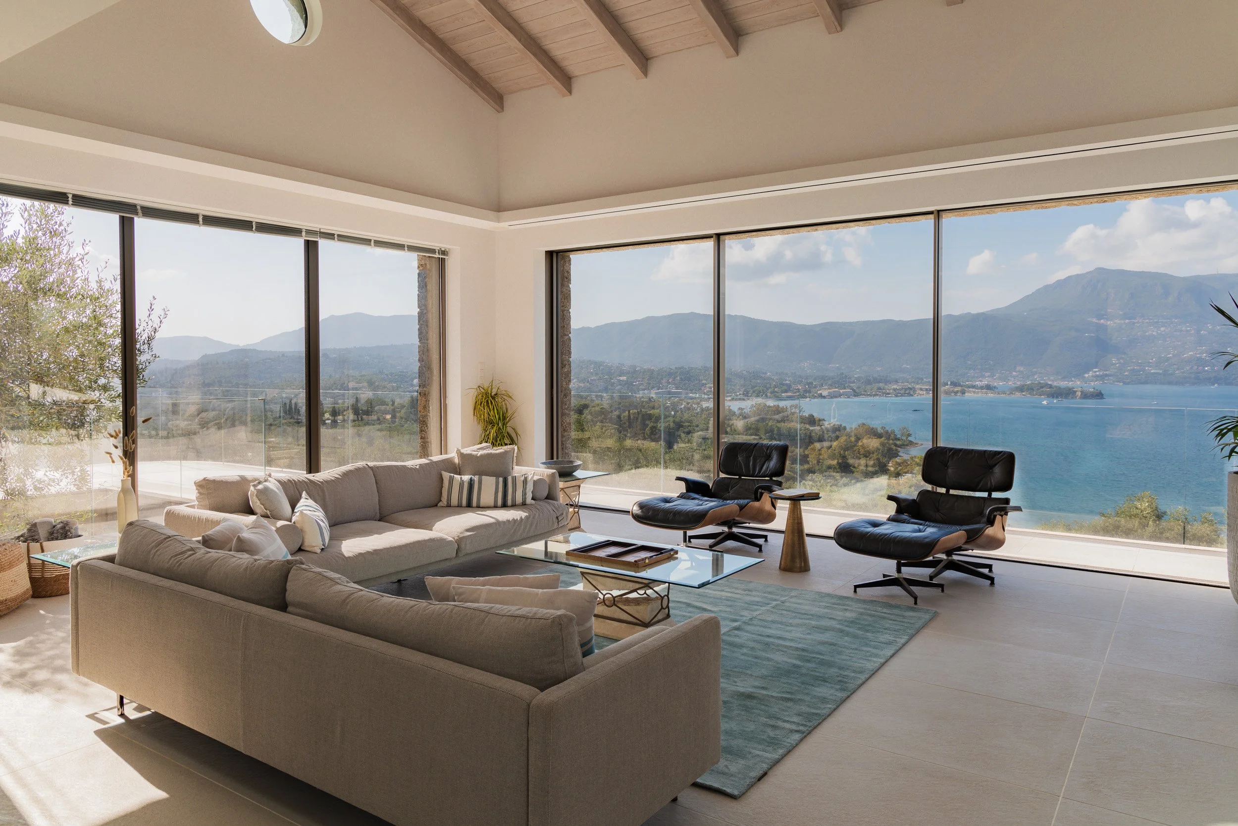 Modern living room with large floor-to-ceiling windows overlooking a lake and mountains, featuring sofas, armchairs, a glass coffee table, and indoor plants.