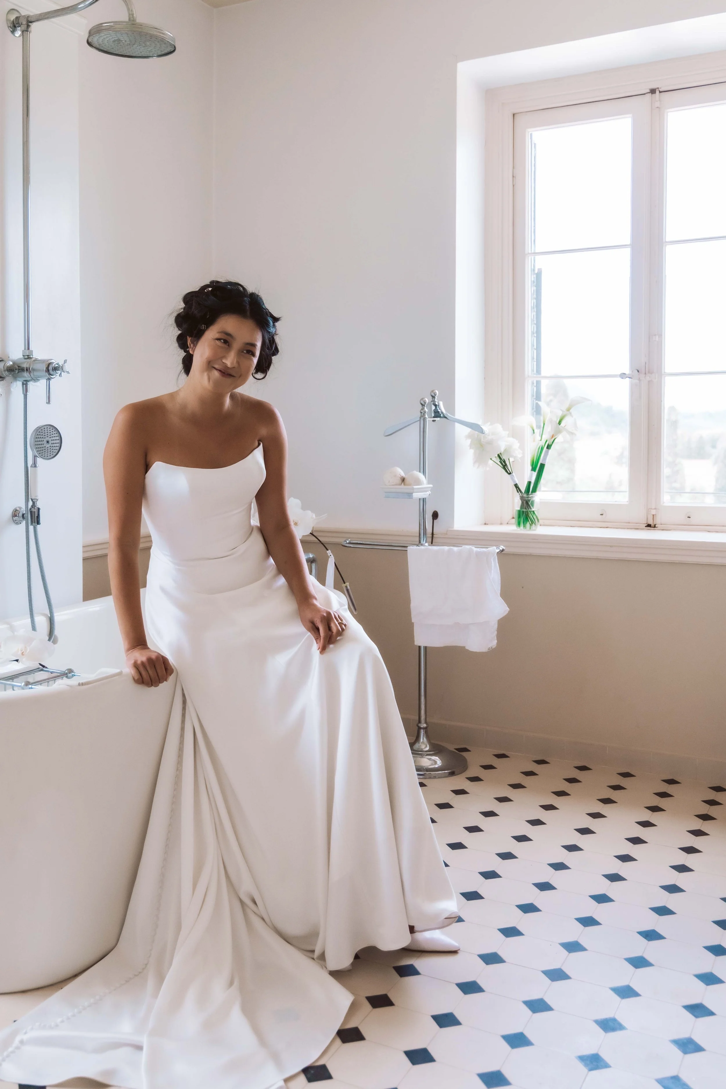 A woman in a white wedding dress sitting on the edge of a bathtub in a bright, minimalist bathroom with black and white tiled floor, large window, and a vase of white flowers.