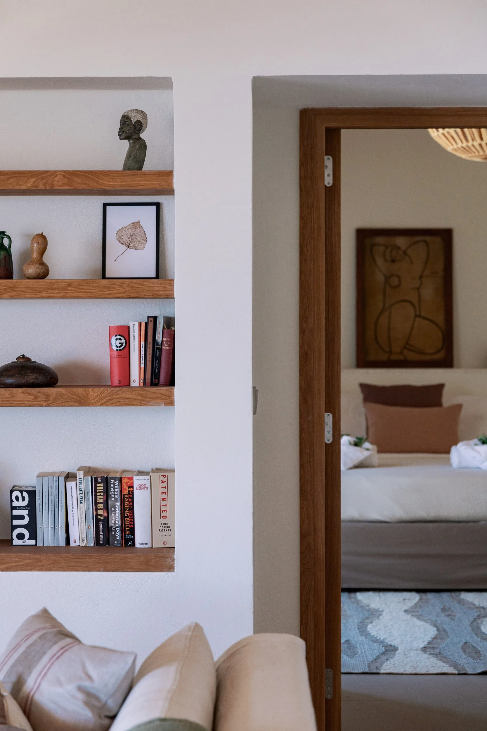 Living room with bookshelf on the left, decorative items on the shelves, and an open doorway leading to a bedroom with bed and framed artwork.