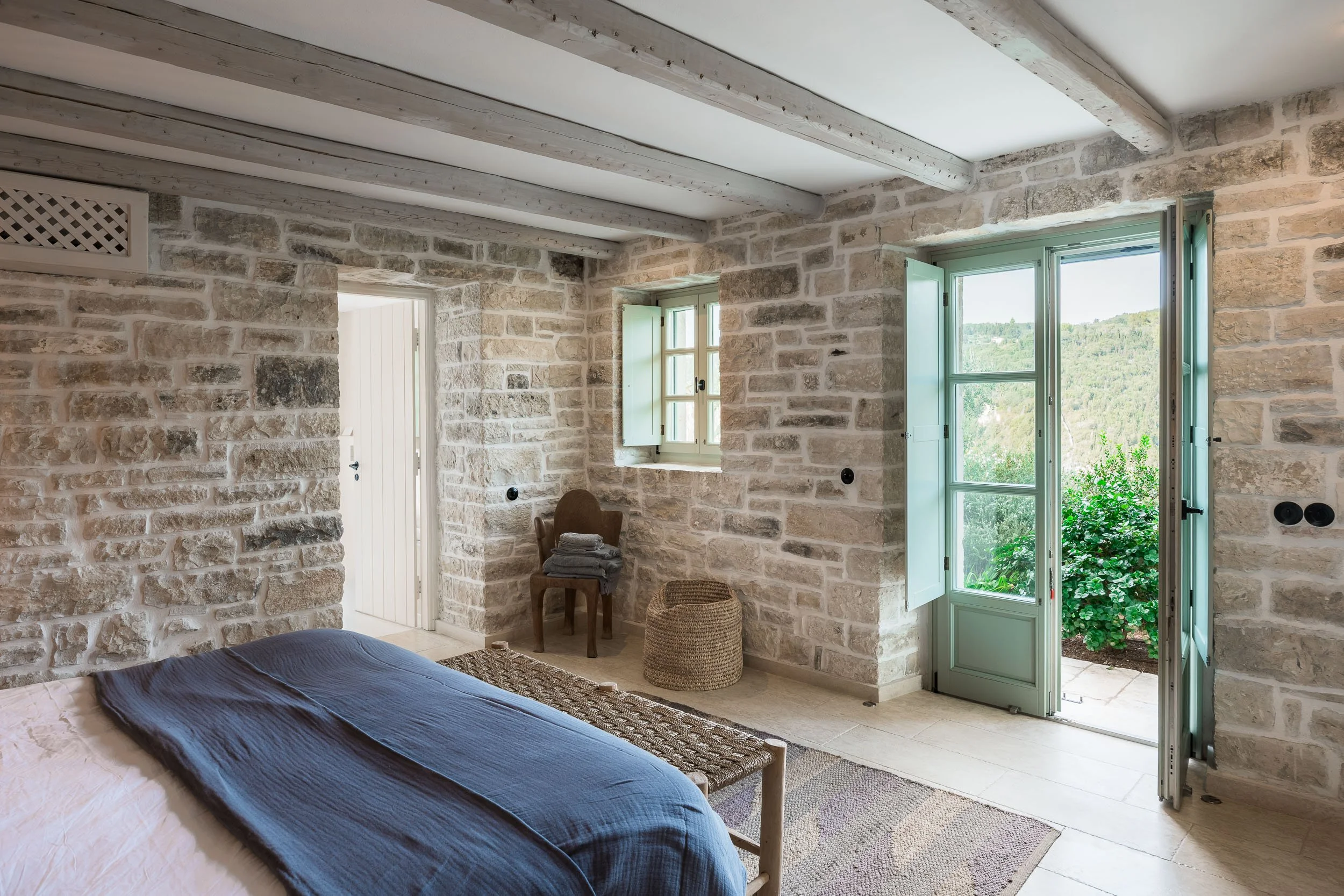 Bedroom with stone walls, an open door leading to a green outdoor view, and a bed with dark blue bedding.