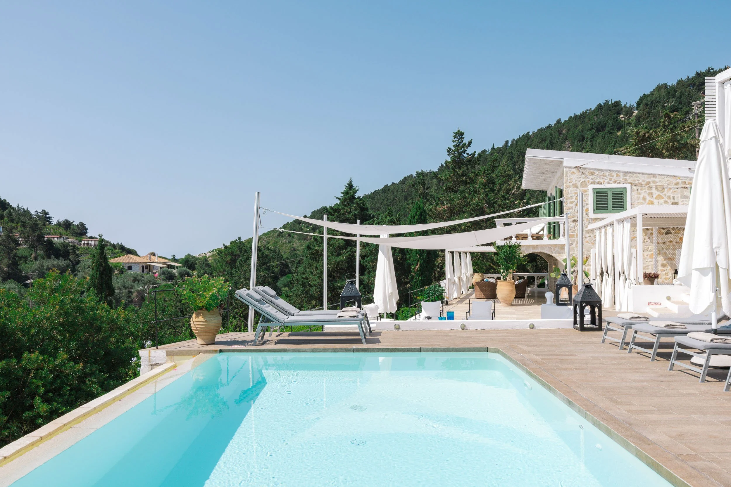 Resort-style outdoor pool area with lounge chairs, umbrellas, and a stone house, surrounded by green hills under a clear blue sky.