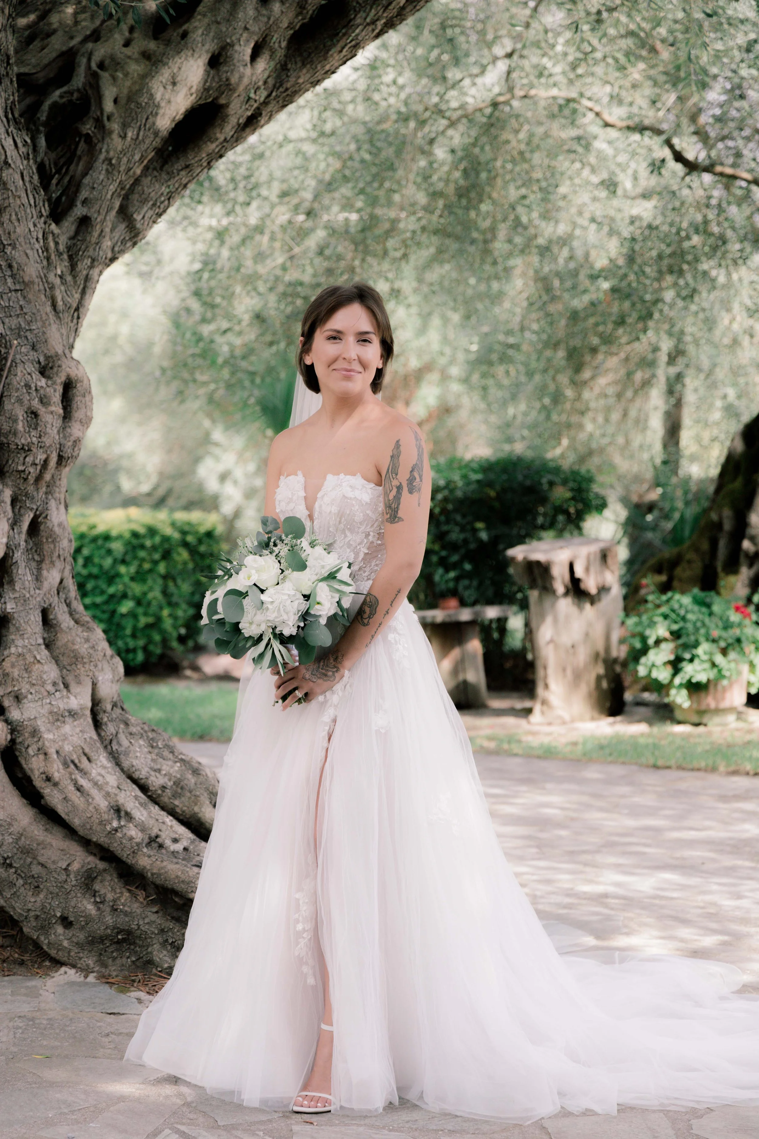 A smiling bride in a white wedding dress holding a bouquet of white flowers, standing outdoors near a large tree with lush greenery in the background.