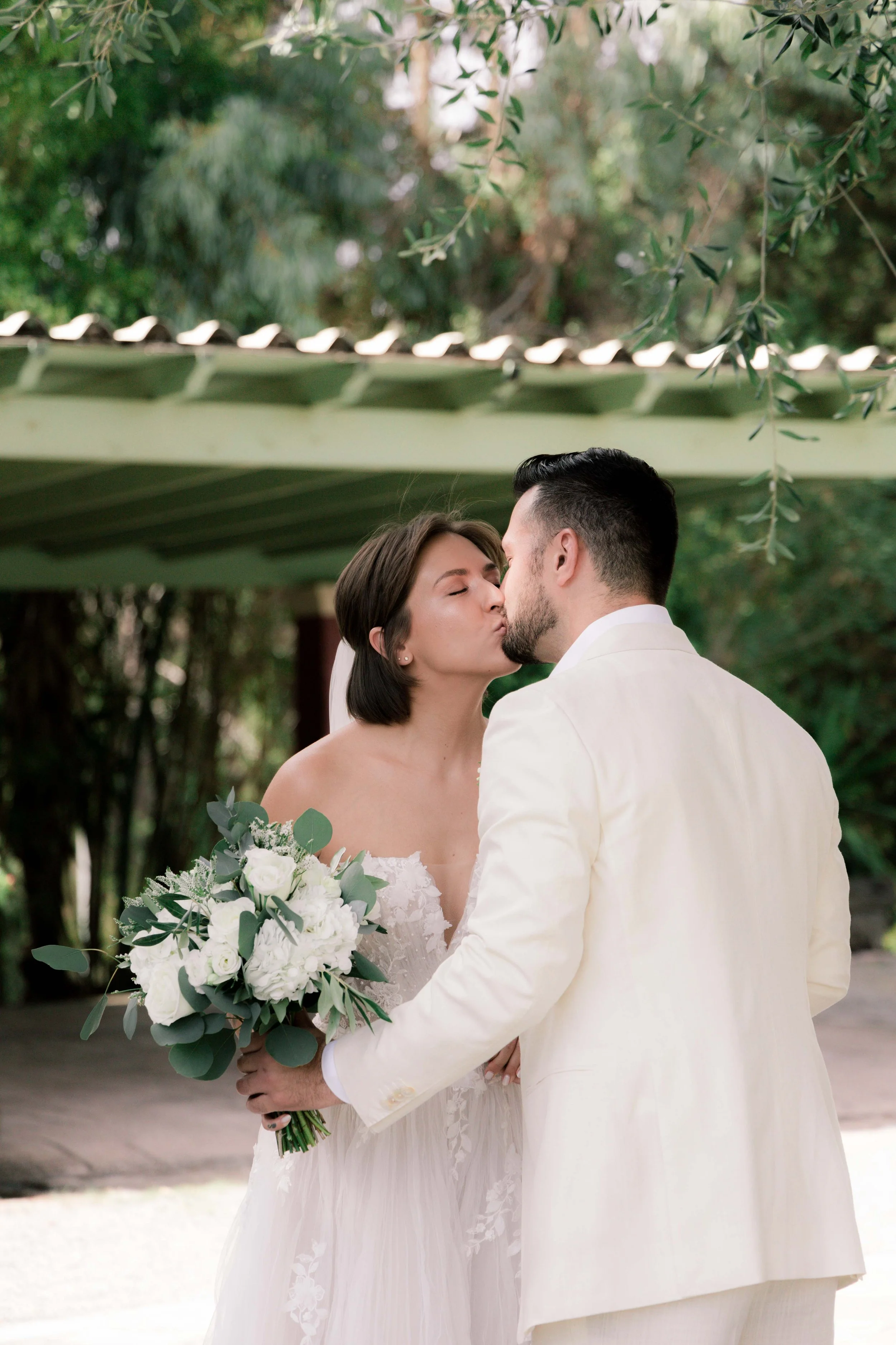 A bride and groom kissing outdoors, the bride holding a bouquet of white flowers and greenery, in front of greenery and a green structure.