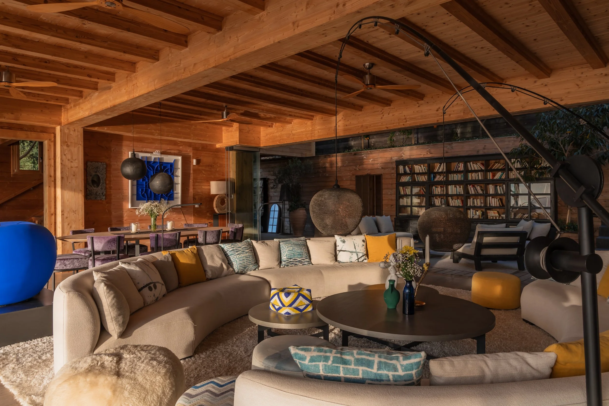 Spacious living room with wooden beams, large sectional sofa with colorful cushions, coffee tables with vases and flowers, hanging light fixtures, bookshelf, and a telescope.
