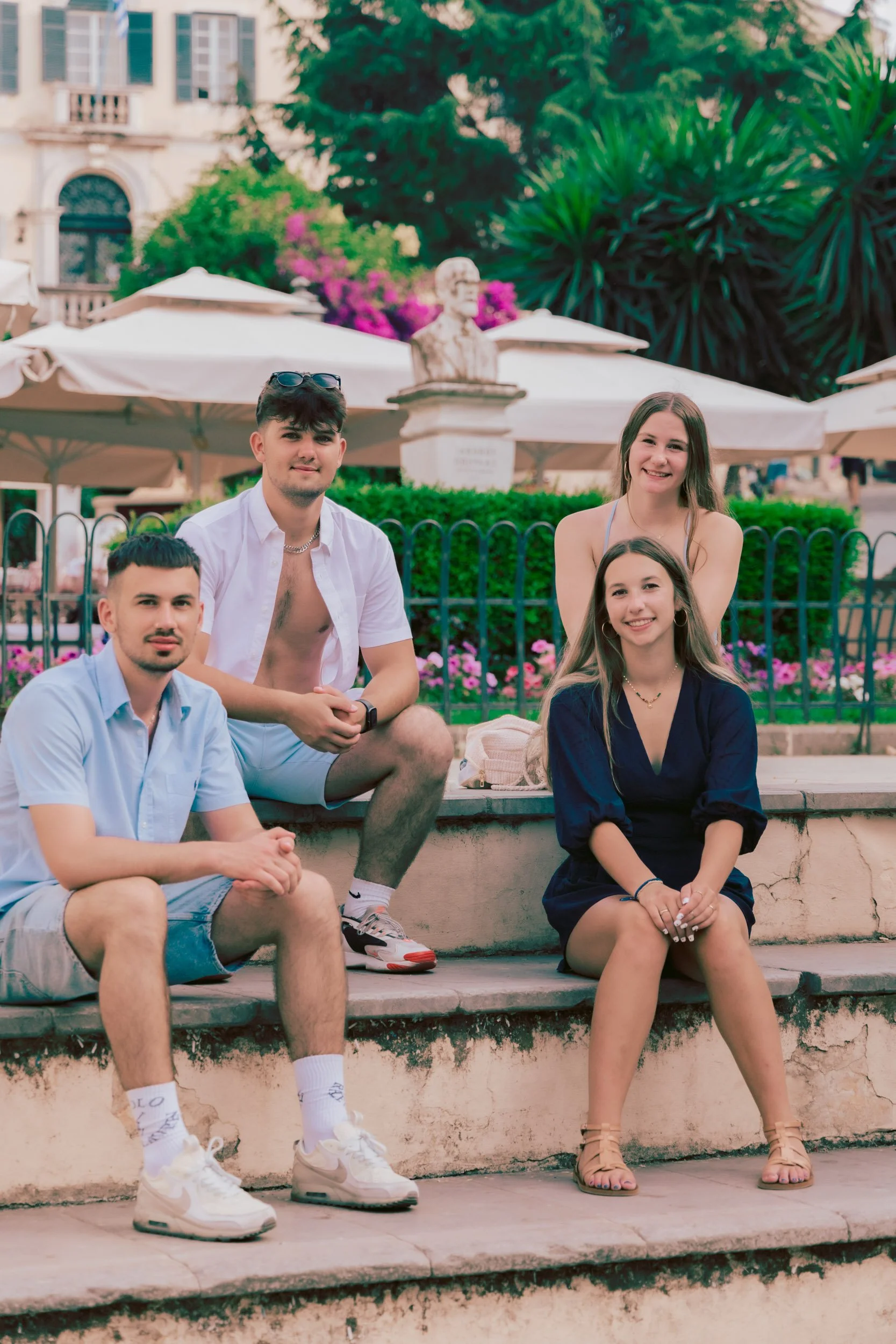 Group of four young adults sitting on city steps, with a park and umbrella-covered tables in the background.