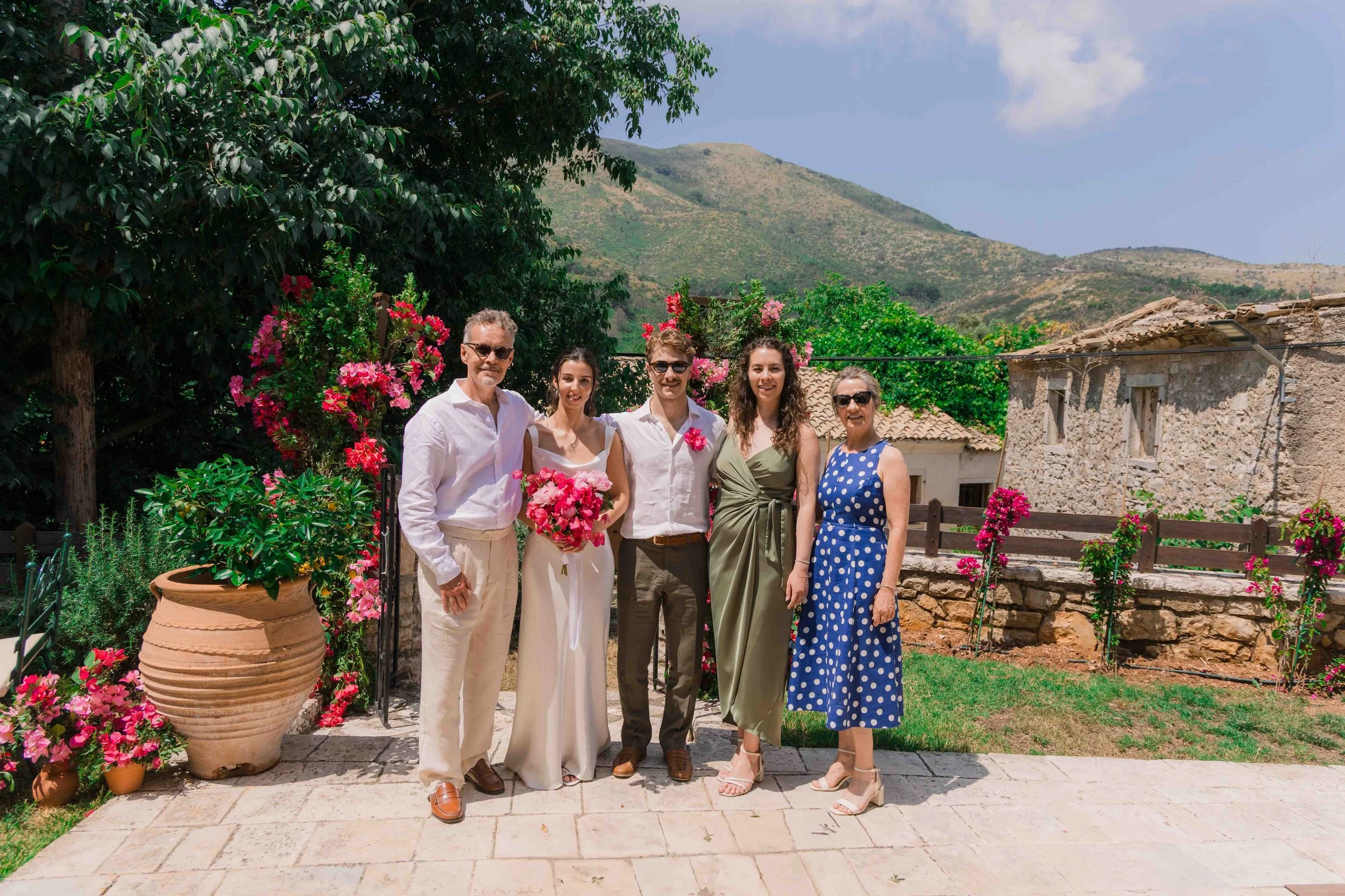 Group of six people standing outdoors in a garden, with hills and a house in the background. The group includes two men and four women dressed in casual and semi-formal attire, some holding flowers, on a sunny day.