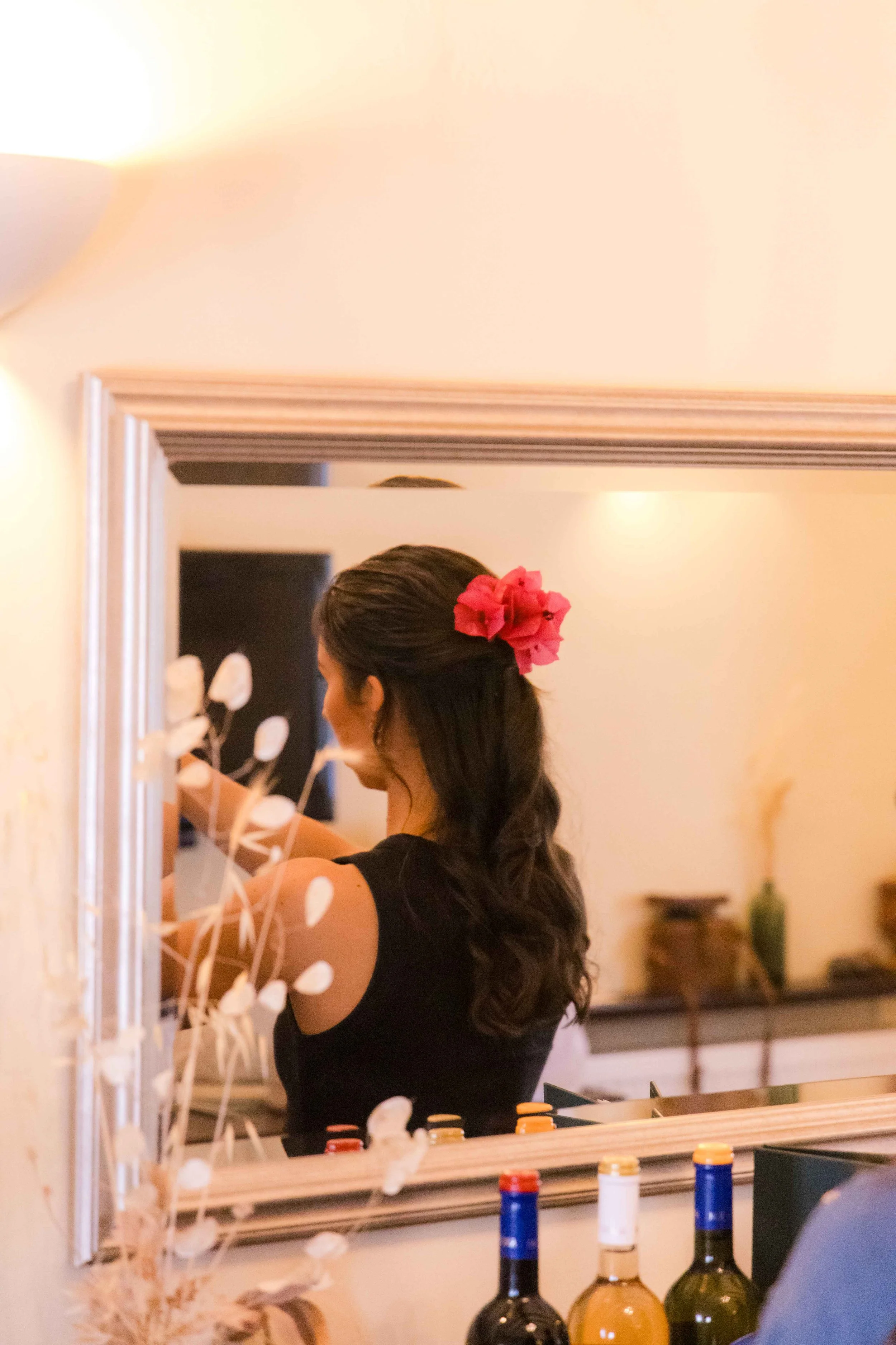 Woman with dark hair and a pink flower clip, seen from the side, in front of a mirror, preparing or adjusting her hair.