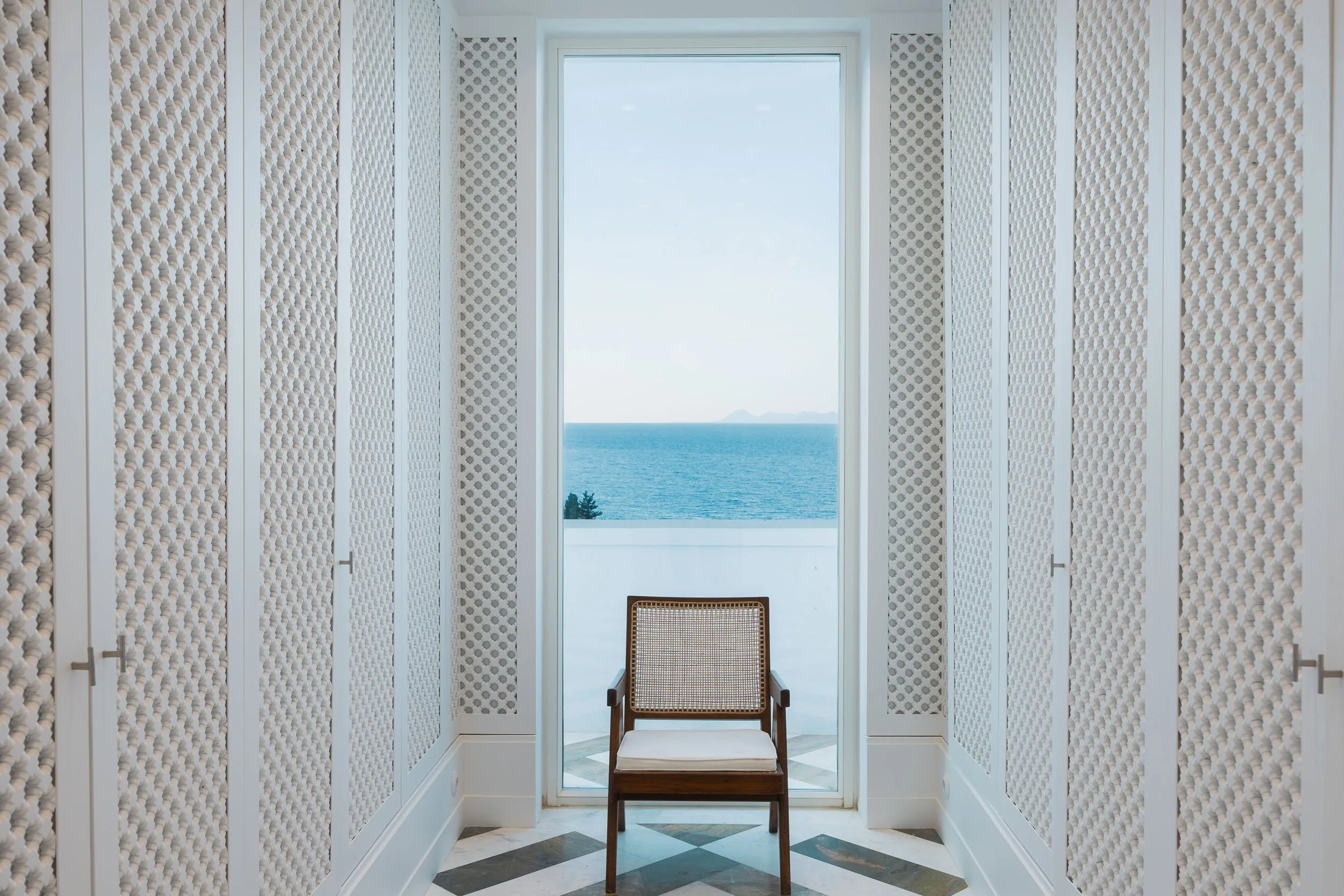 Interior of a room with white lattice walls, a wooden chair with a woven back and cushion, and a large glass door leading to a balcony with an ocean view.