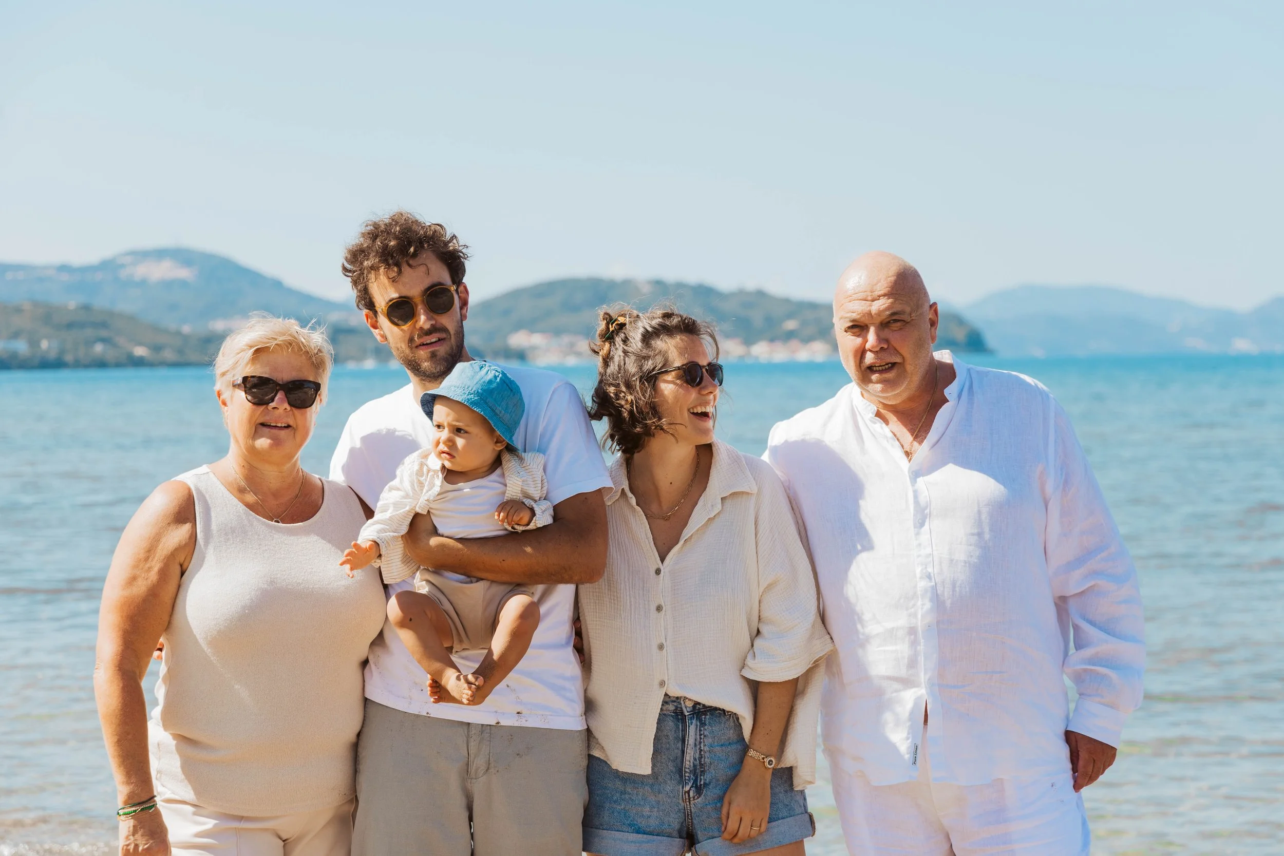 A group of six people, including children, standing on a beach with water and hills in the background on a sunny day.