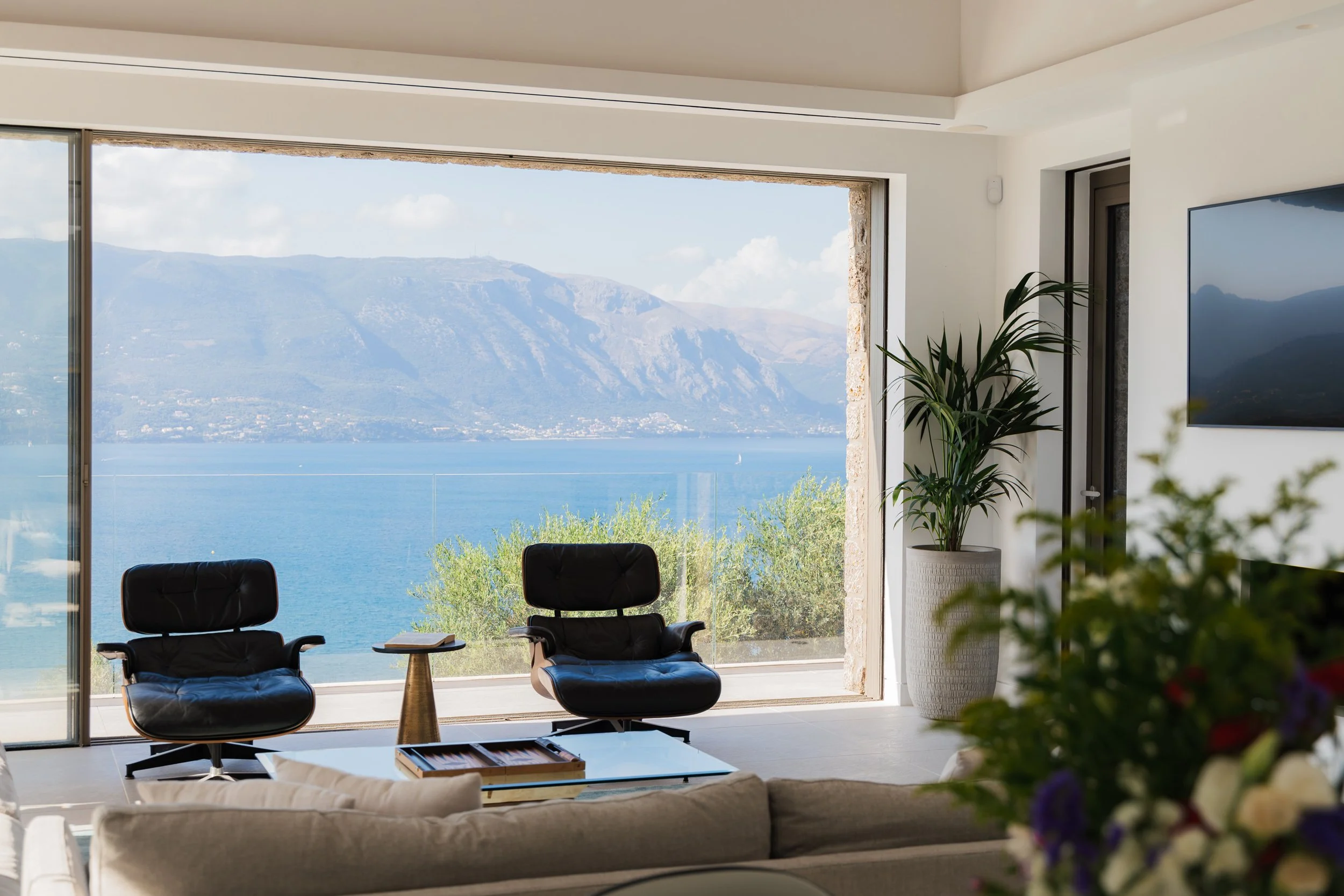 Living room with large window overlooking a lake and mountains, two black chairs, a small side table, a potted plant, and a flat-screen TV.