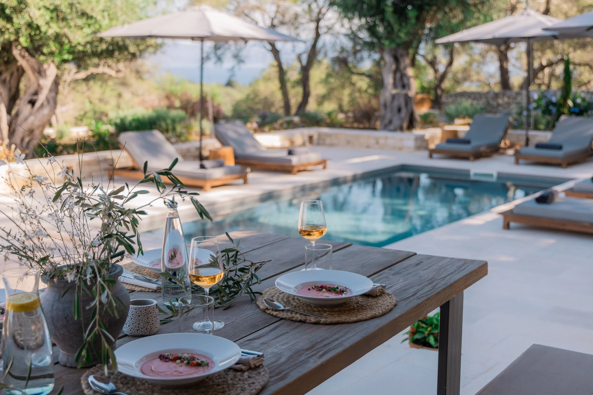 Outdoor dining setup near a swimming pool with plates of pink soup, wine glasses, and decorative greenery, surrounded by lounge chairs and umbrellas with trees in the background.