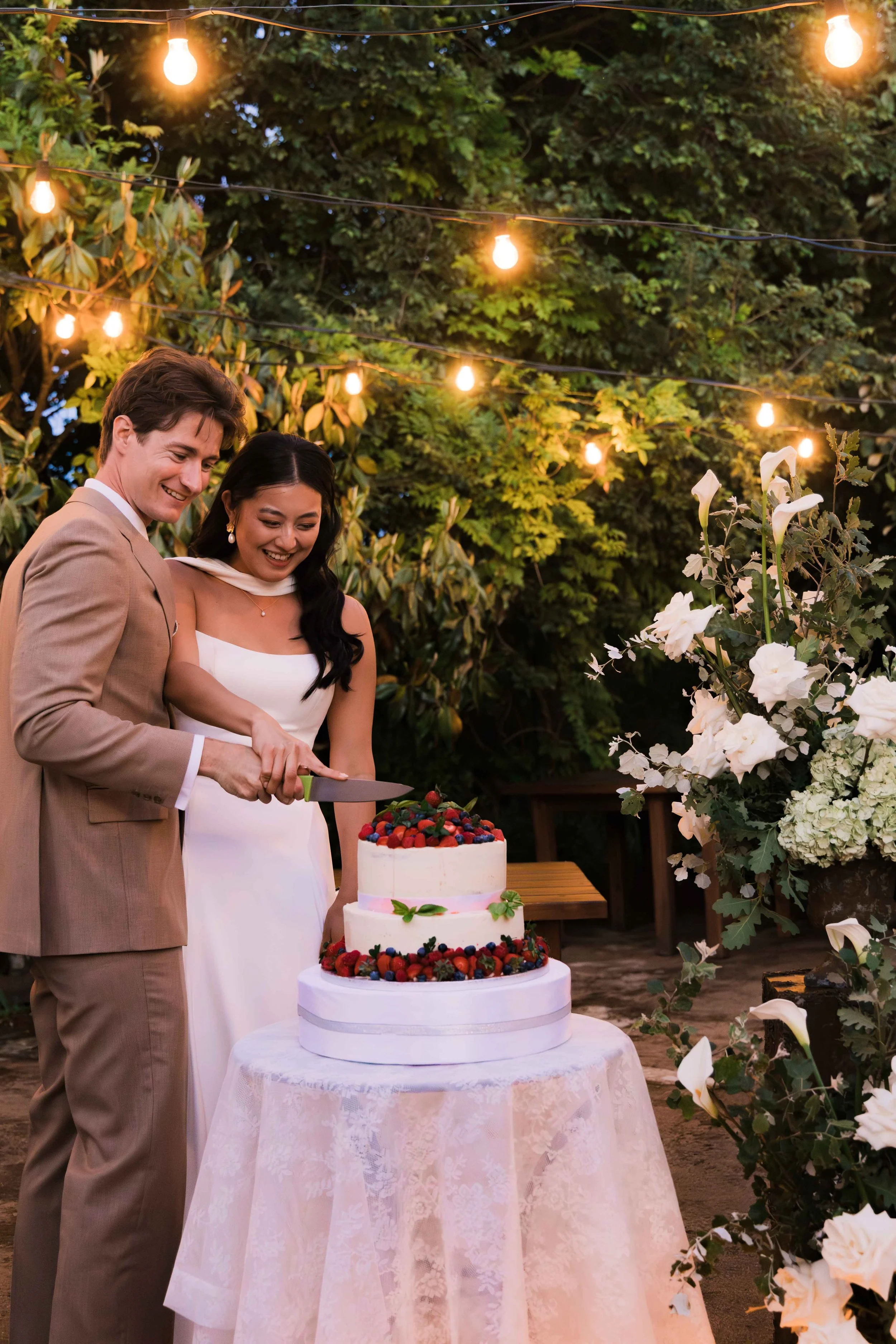 A couple celebrating a wedding outdoors, cutting a three-tiered wedding cake decorated with berries, under string lights surrounded by greenery and white flowers.