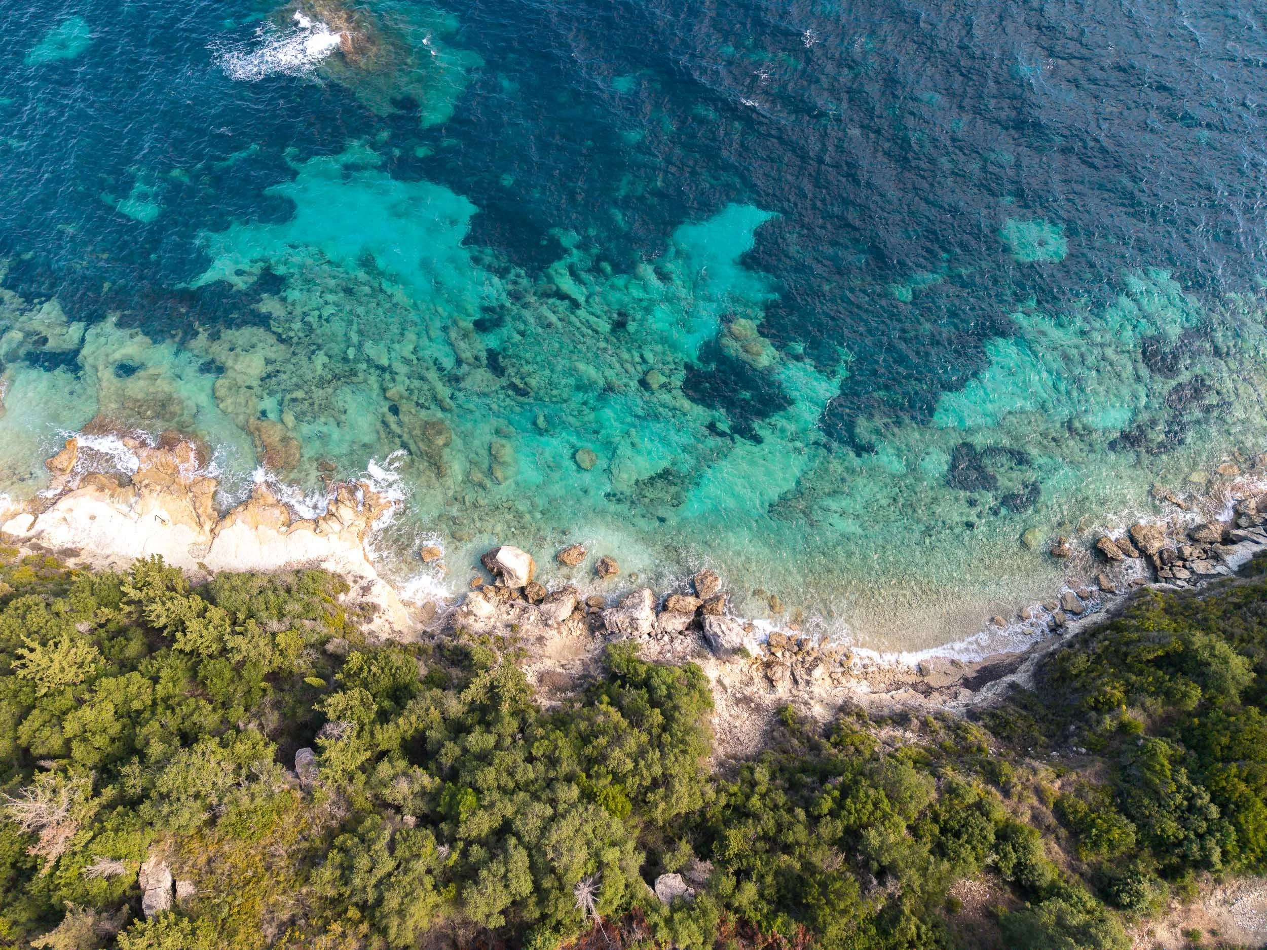 Aerial view of ocean coastline with shallow turquoise waters, rocky shoreline, and green forested area.