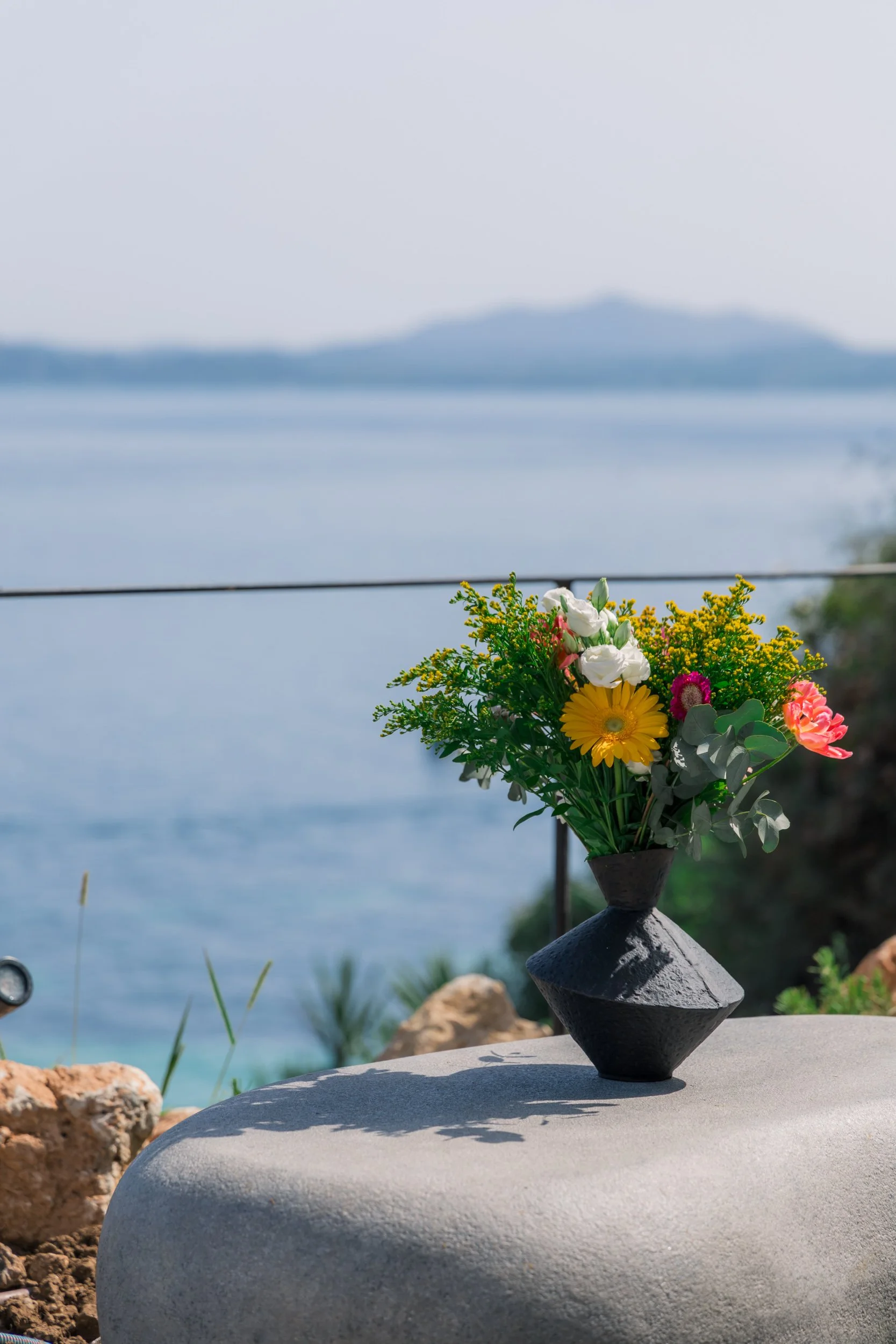 A black vase with colorful flowers, including yellow, pink, and white, sits on a smooth, light-colored surface outdoors, with a body of water and distantlandform in the background.
