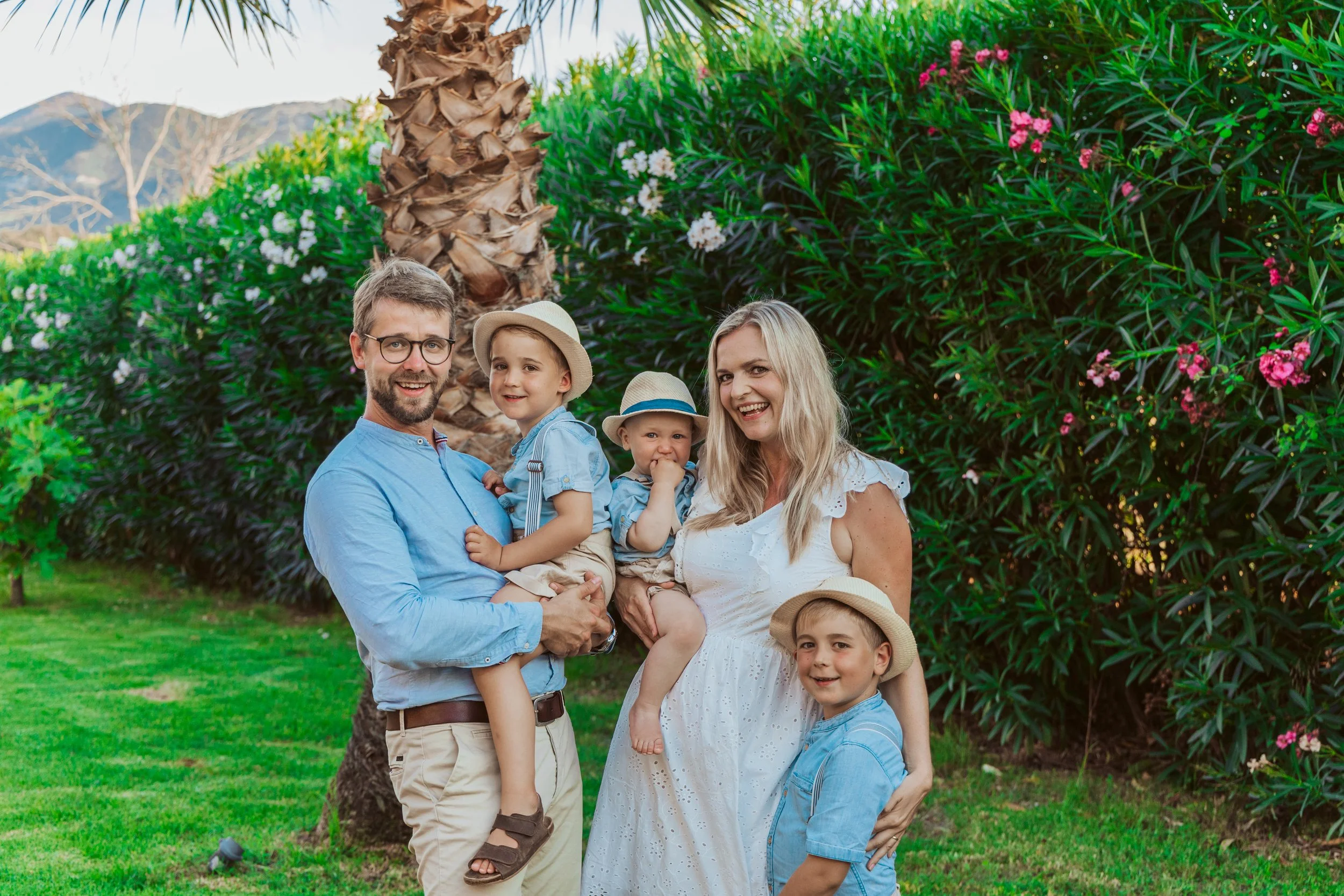 A family of six smiling and standing outdoors near a palm tree and flowering bushes on a sunny day.