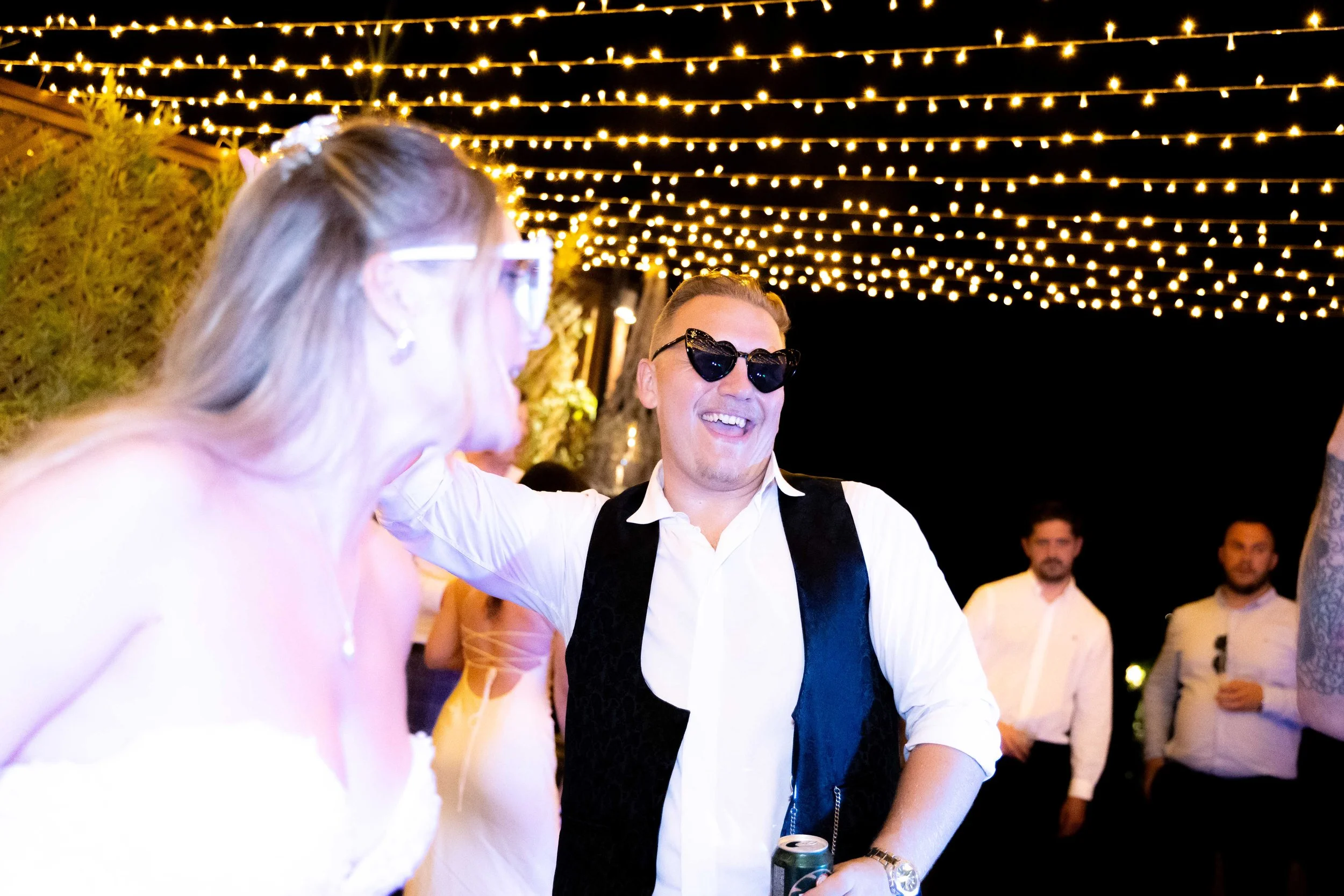 People dancing and enjoying a party under string lights at night, with a man wearing sunglasses and a vest smiling.