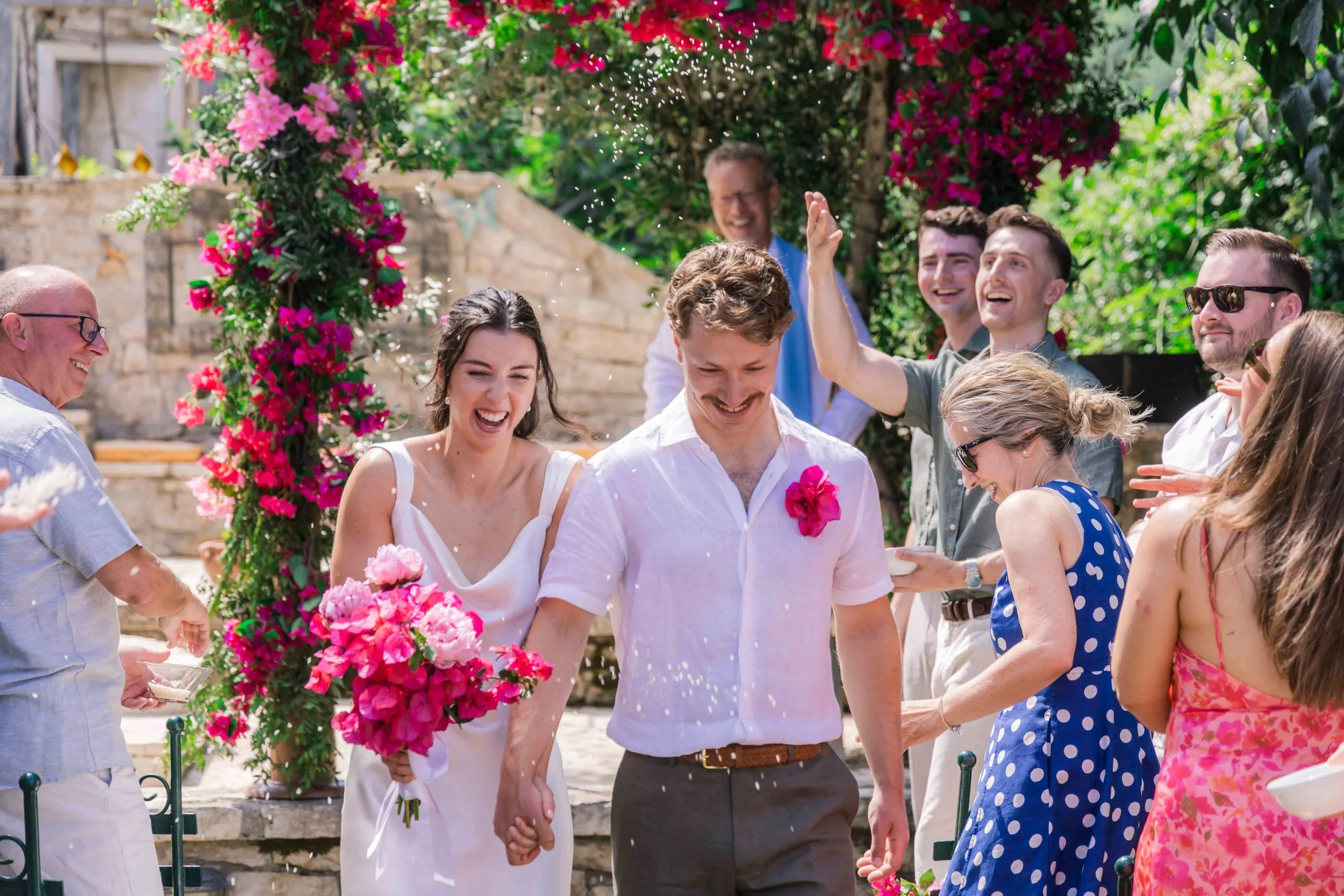 A newlywed couple holding hands and smiling as friends celebrate outdoors near a floral archway with pink blooms, under a sunny sky.