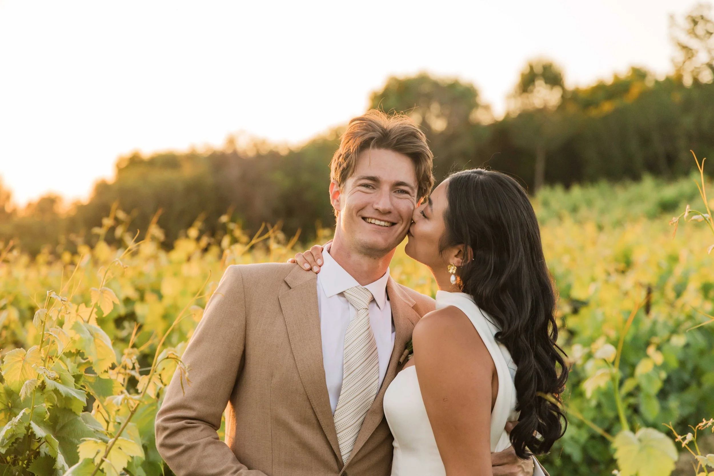 A couple in wedding attire sharing a kiss and smiling outdoors in a vineyard during sunset.