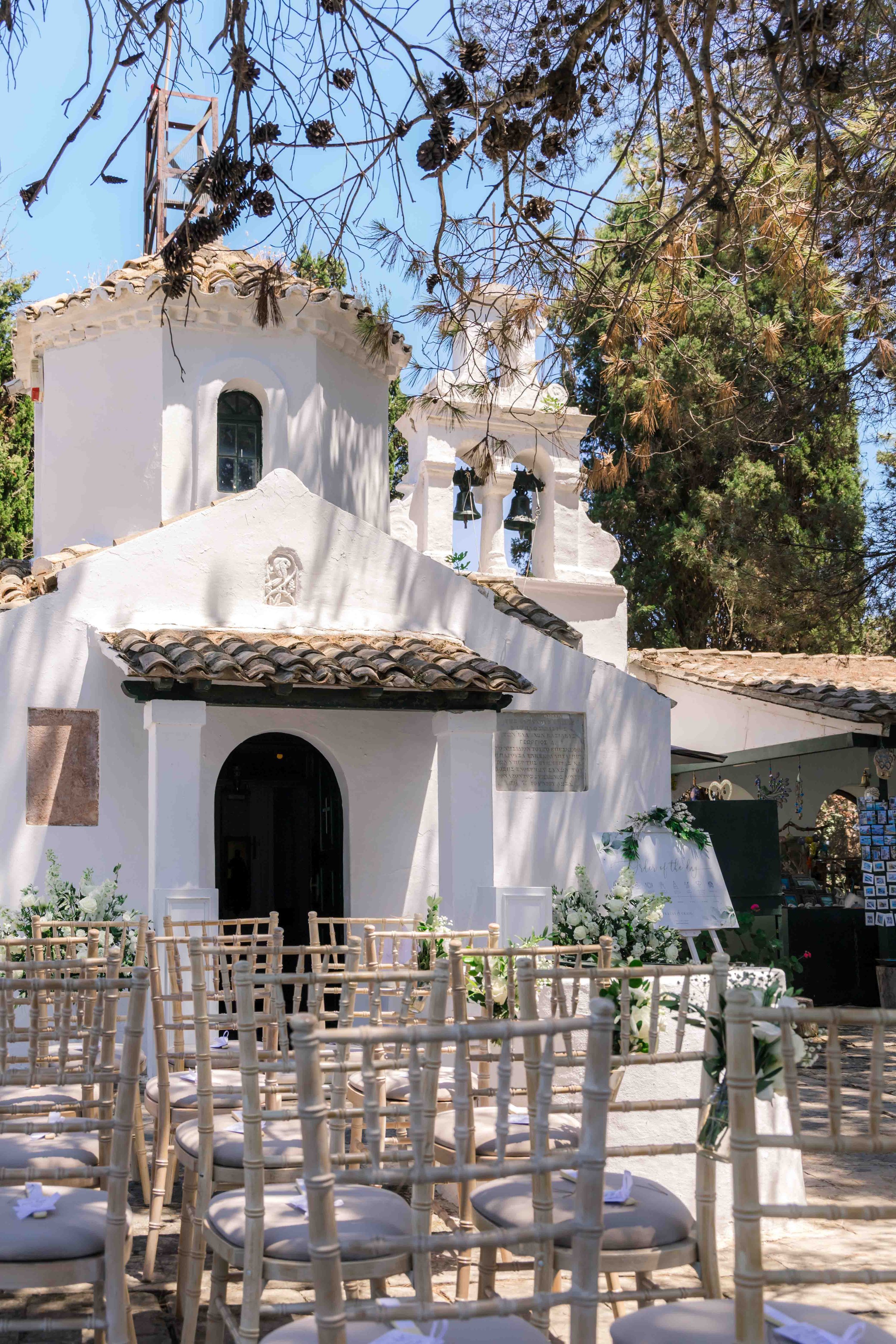 White chapel with outdoor seating arranged with wooden chairs and white cushions, decorated with white flowers and greenery, under trees with pinecones, likely set up for a wedding or ceremony.