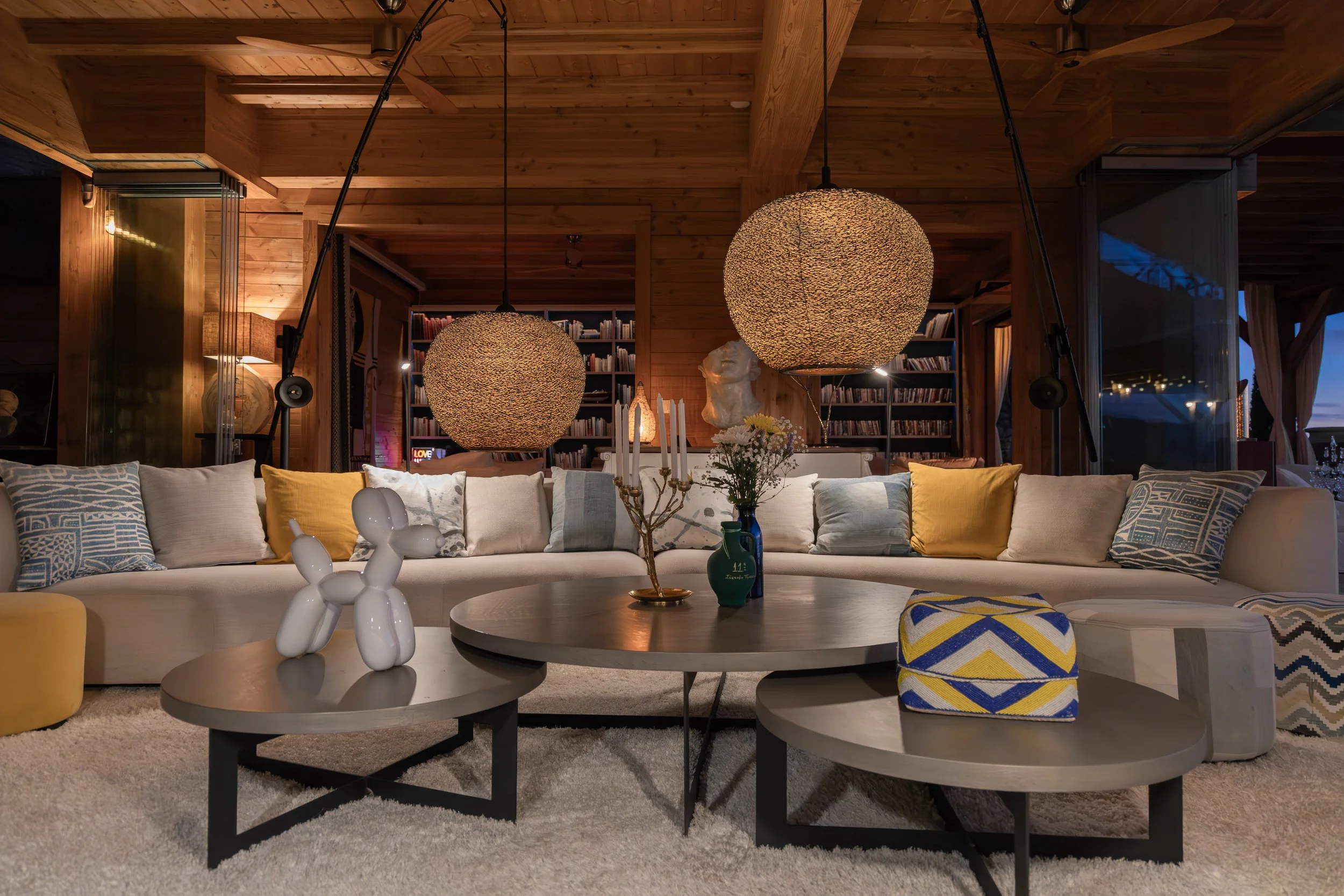 Living room with a large beige sectional sofa decorated with yellow and patterned pillows, wooden ceiling with two large round woven pendant lights, coffee tables with vases, and built-in bookshelves in the background.