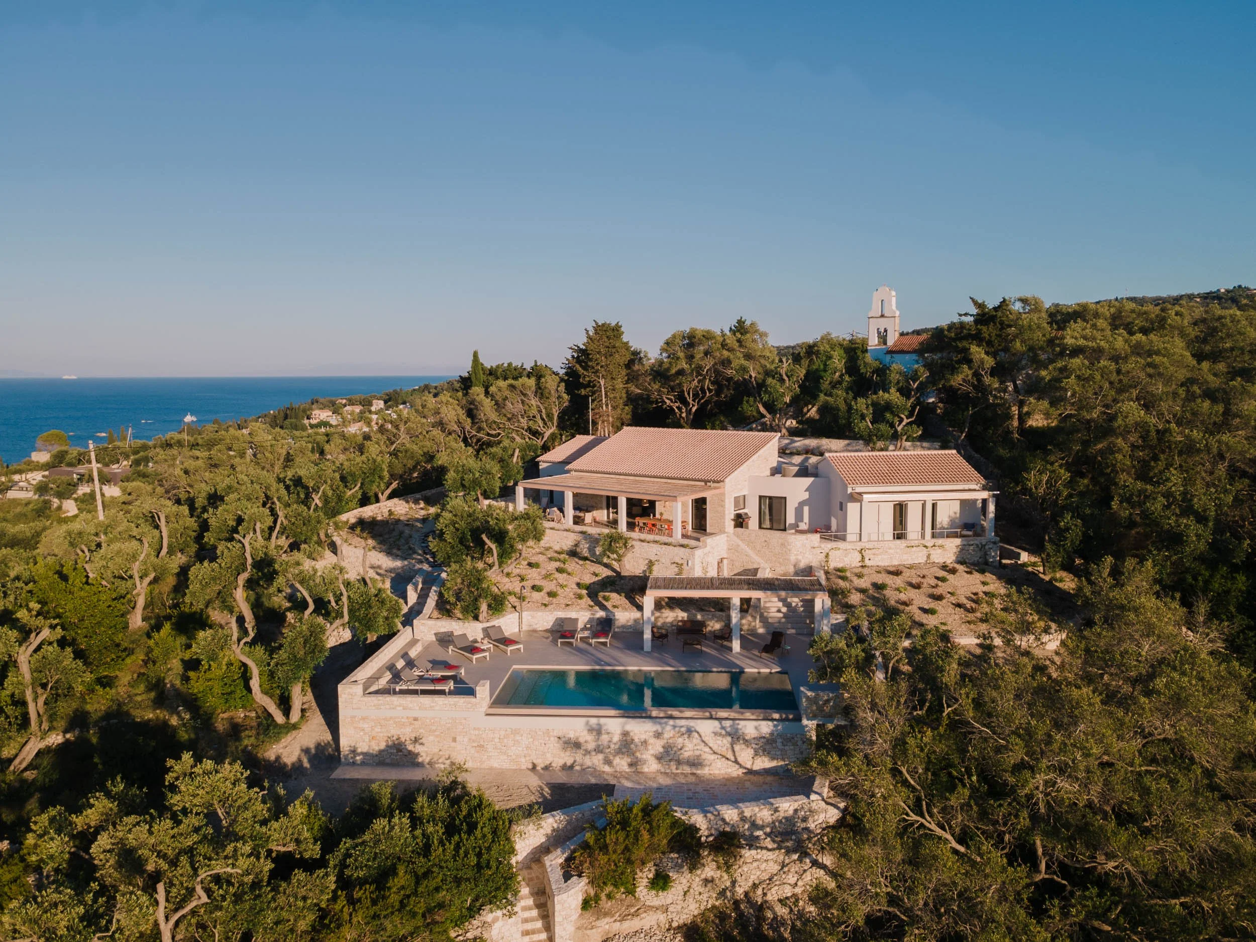 Aerial view of a luxurious house with a swimming pool on a hillside, surrounded by trees, overlooking the ocean under a clear blue sky.