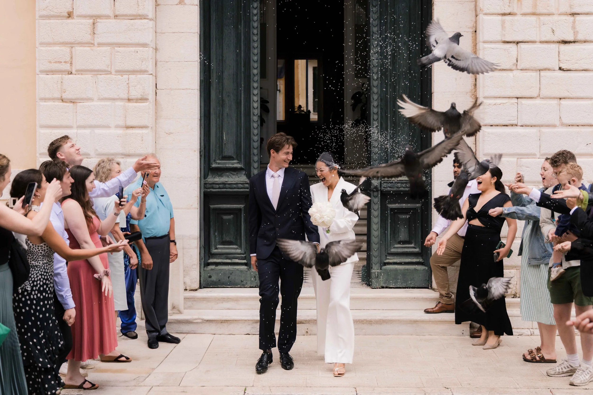 A joyful bride and groom walking out of a building with open green double doors, surrounded by guests throwing birdseed and taking photos. The bride is holding a bouquet of white flowers, and the groom is wearing a dark suit. Pigeons are flying aroun