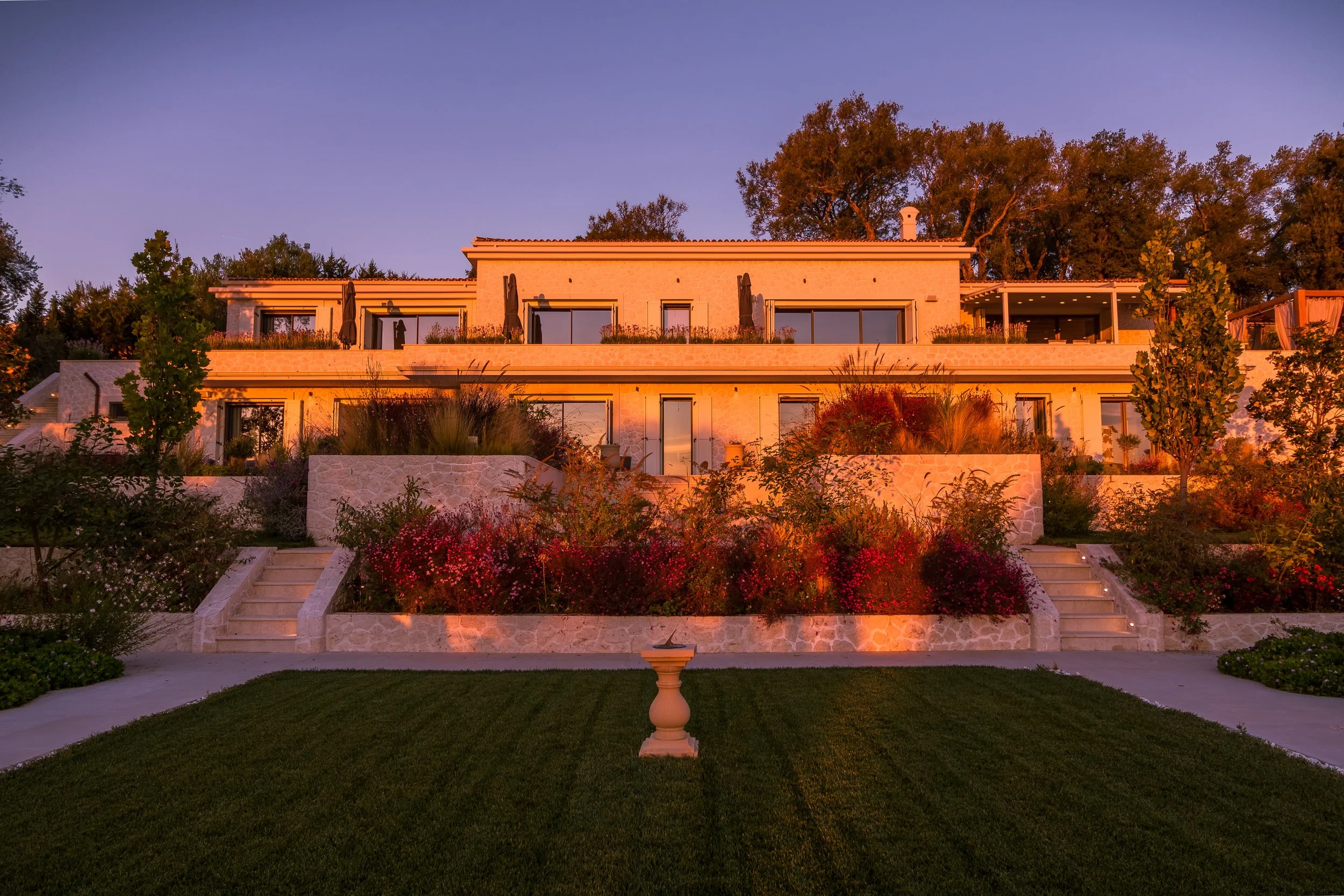A multi-story modern house at sunset, with large windows, surrounded by landscaped garden with trees, shrubs, and flowers, and a green lawn in the foreground.