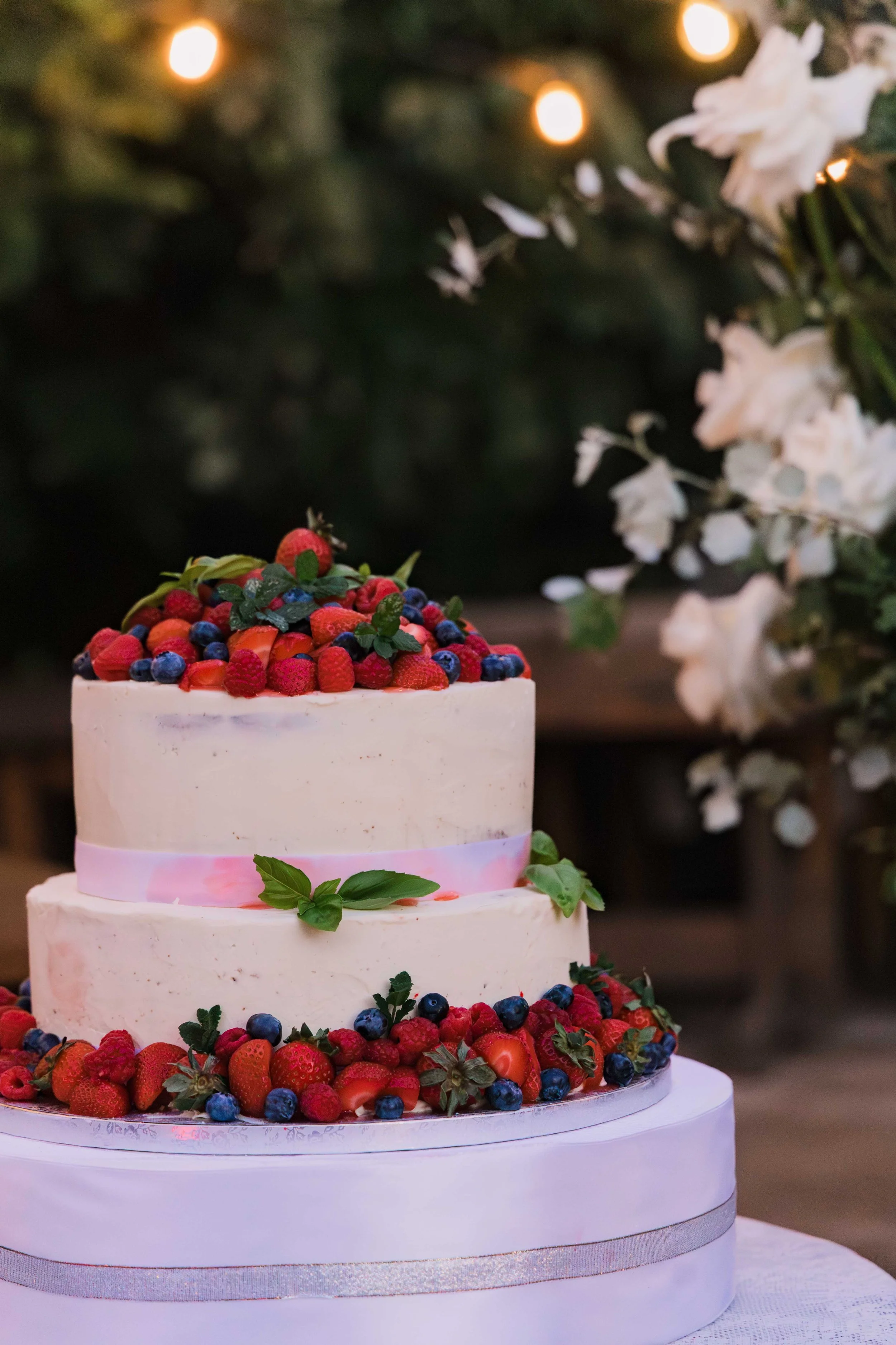 A three-tiered wedding cake decorated with fresh berries and green leaves, with a pink ribbon accent around the middle tier, placed on a white tablecloth with blurred floral and greenery background.