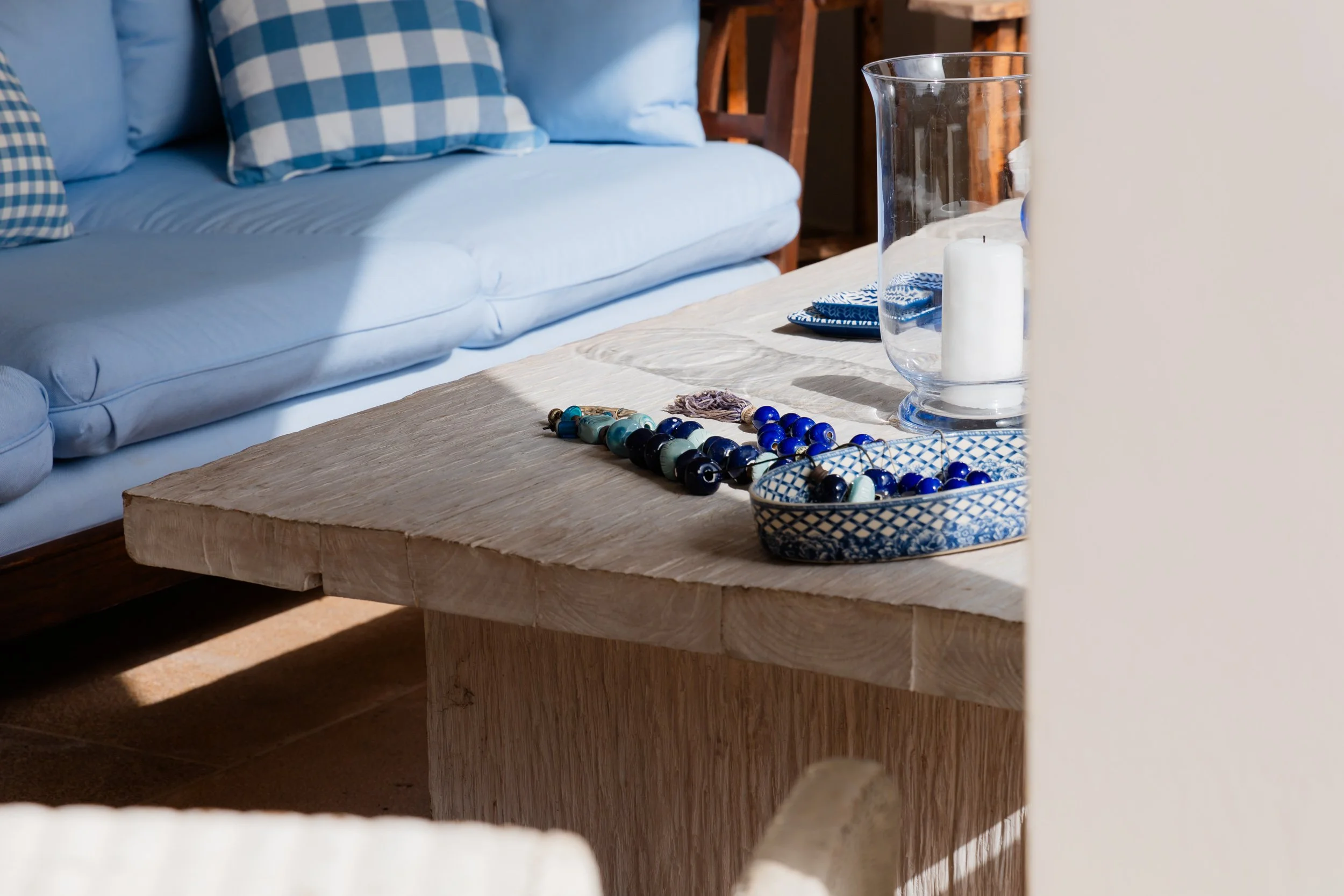A wooden coffee table with blue and turquoise jewelry and beads, a glass hurricane with a white candle, and some blue and white patterned dishware, next to a white sofa with blue checkered pillows.