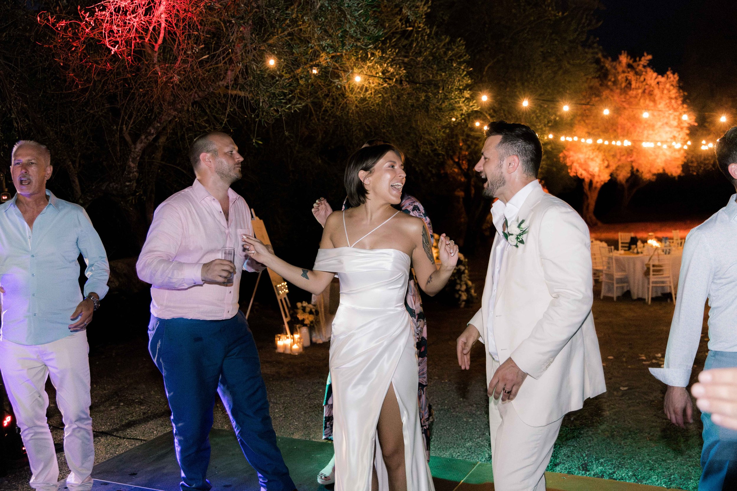 A group of people dancing and celebrating at an outdoor nighttime event, possibly a wedding reception, with string lights, candles, and decorated tables in the background.