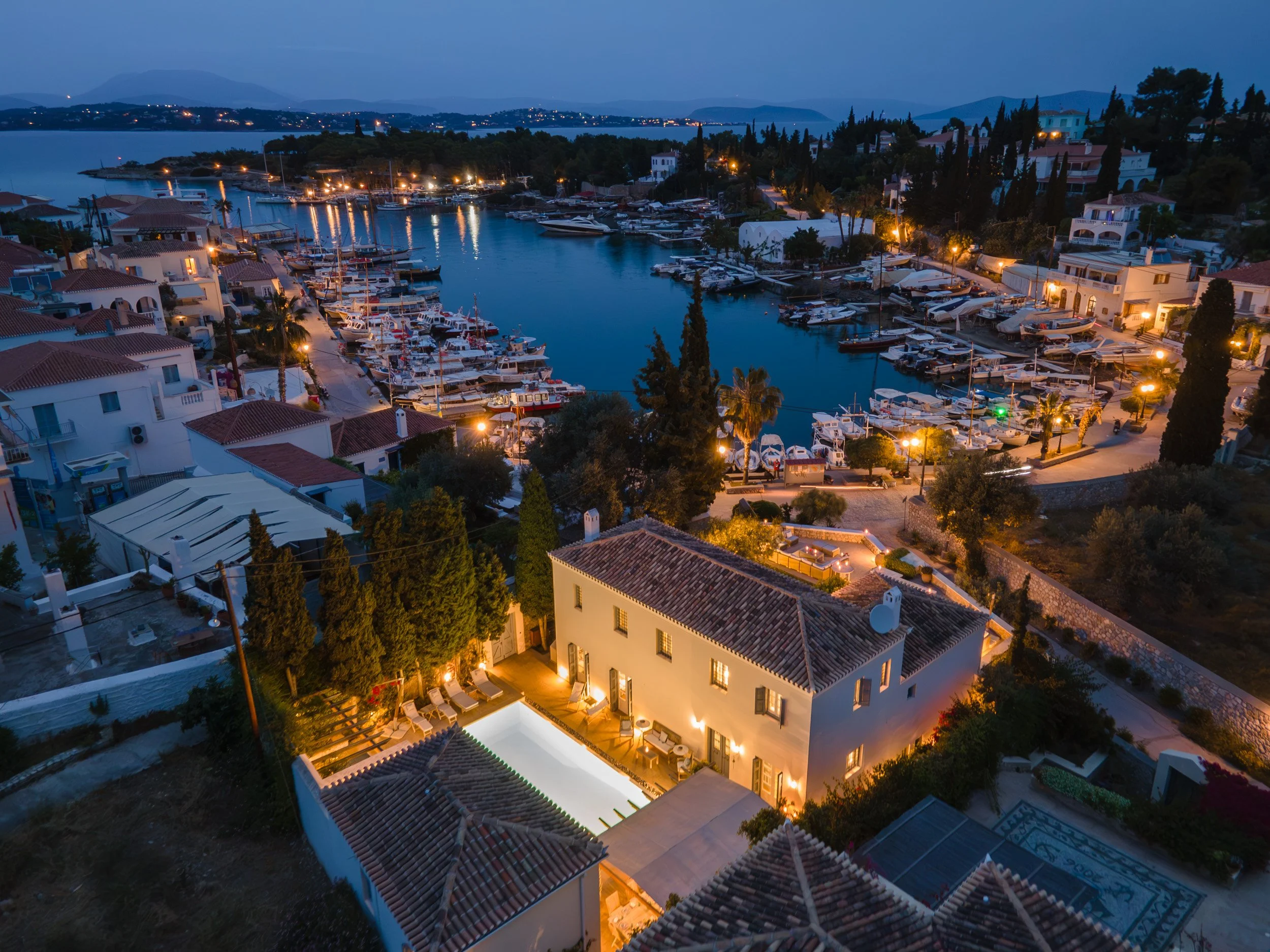 Aerial view of a coastal town at dusk with a marina filled with boats, houses with illuminated windows, and a calm body of water in the background.