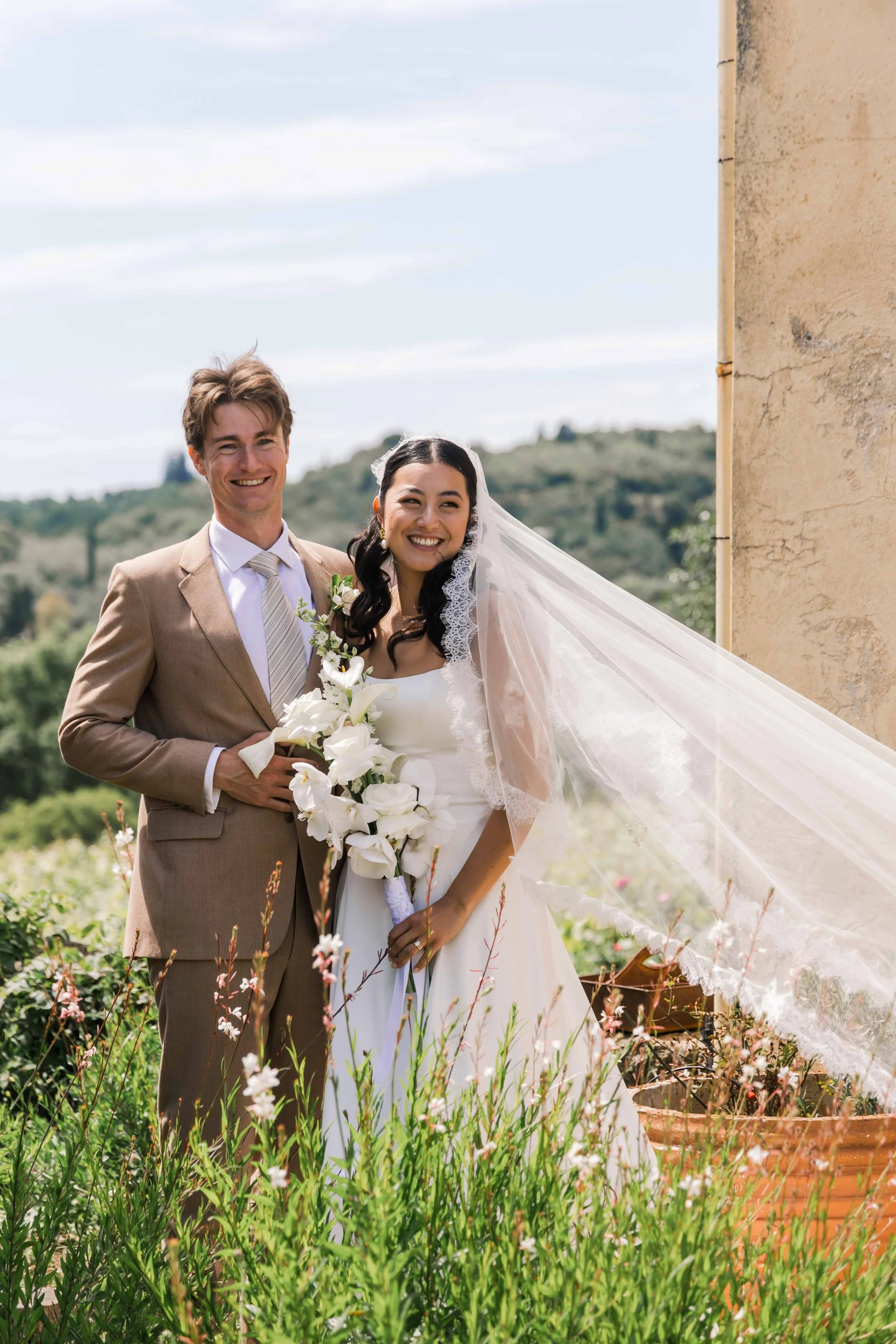 A newlywed couple stands outdoors near a stone wall, smiling and holding a bouquet of white flowers, with green hills and a partly cloudy sky in the background.