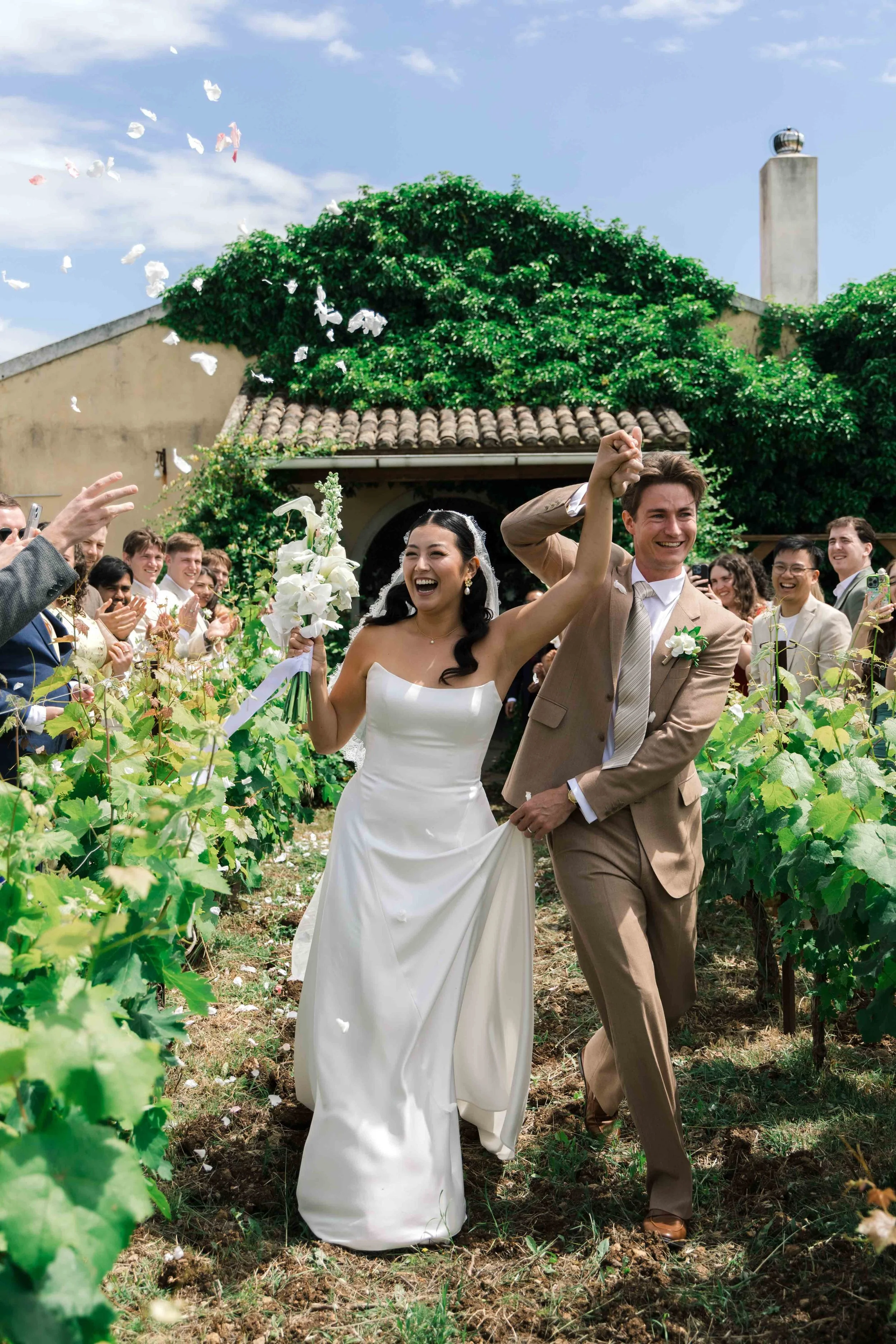 A newlywed couple walking hand-in-hand through a vineyard, smiling and celebrating with friends and family during their wedding ceremony on a sunny day.