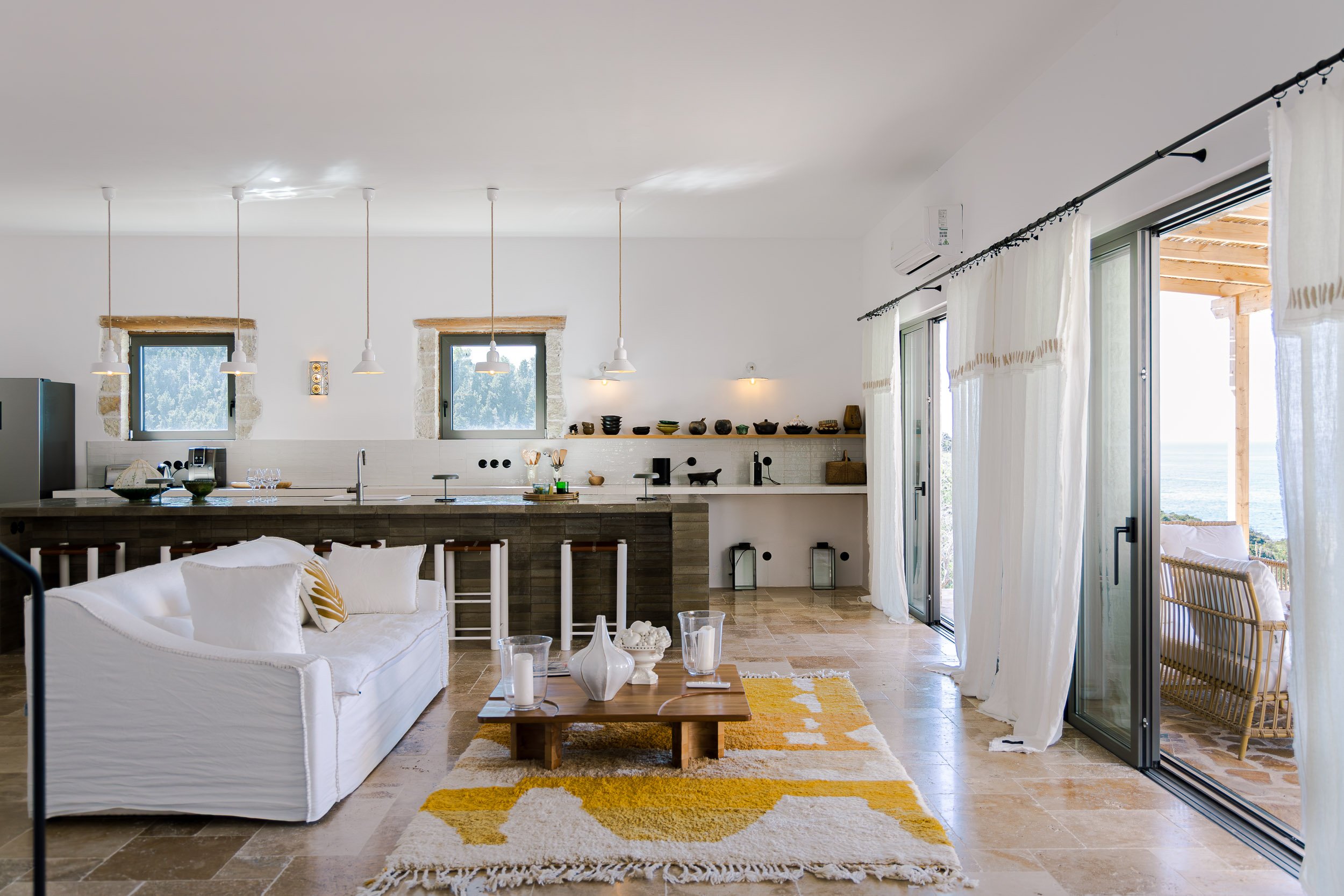 Open-concept living room and kitchen with white walls, pendant lights, a white sofa, and a sliding glass door leading to an outdoor patio with ocean view.