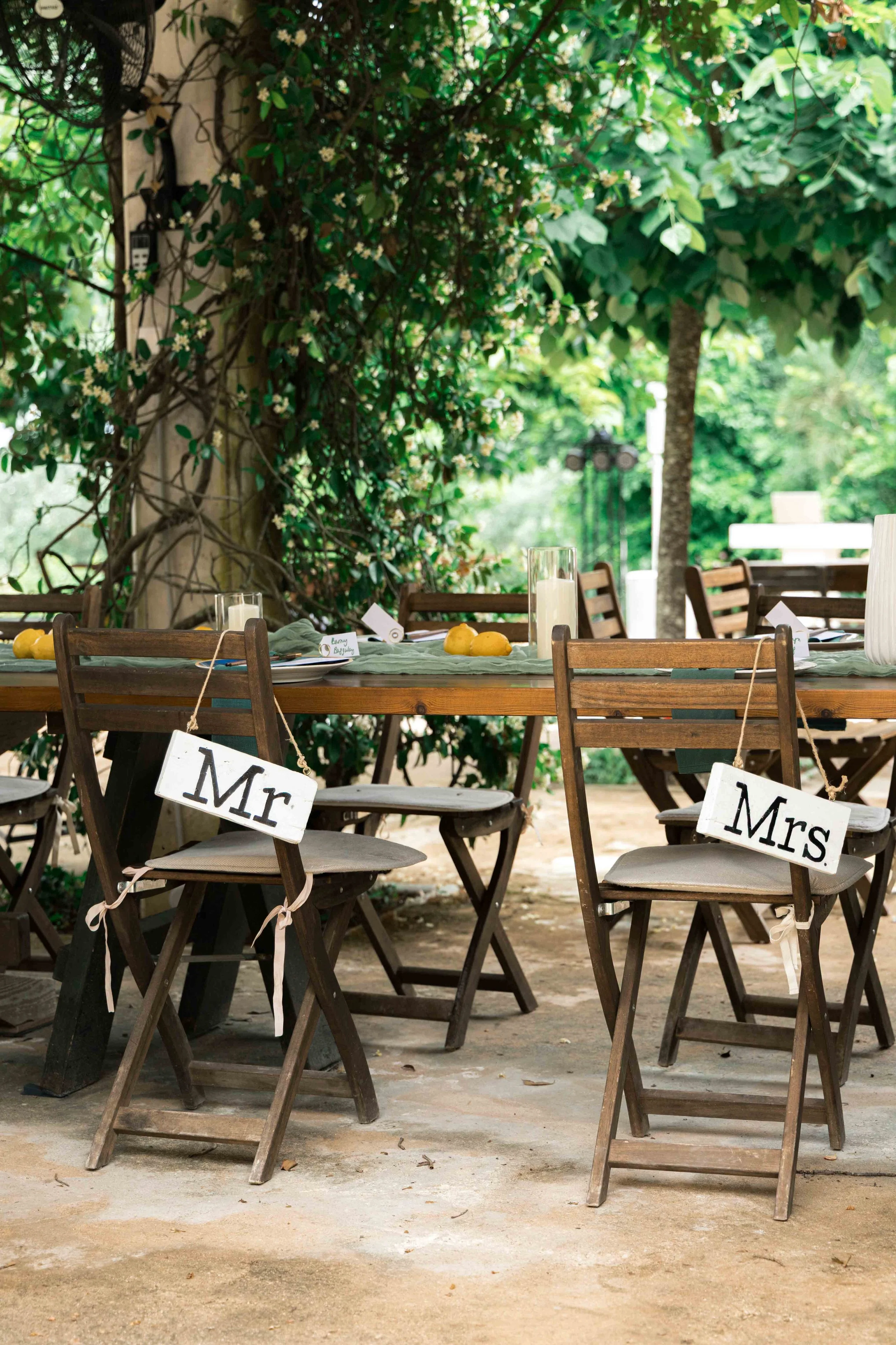 An outdoor table decorated with white candles and yellow lemons, surrounded by chairs with signs labeled 'Mr' and 'Mrs' to indicate seating for a couple, set in a garden with lush green foliage.