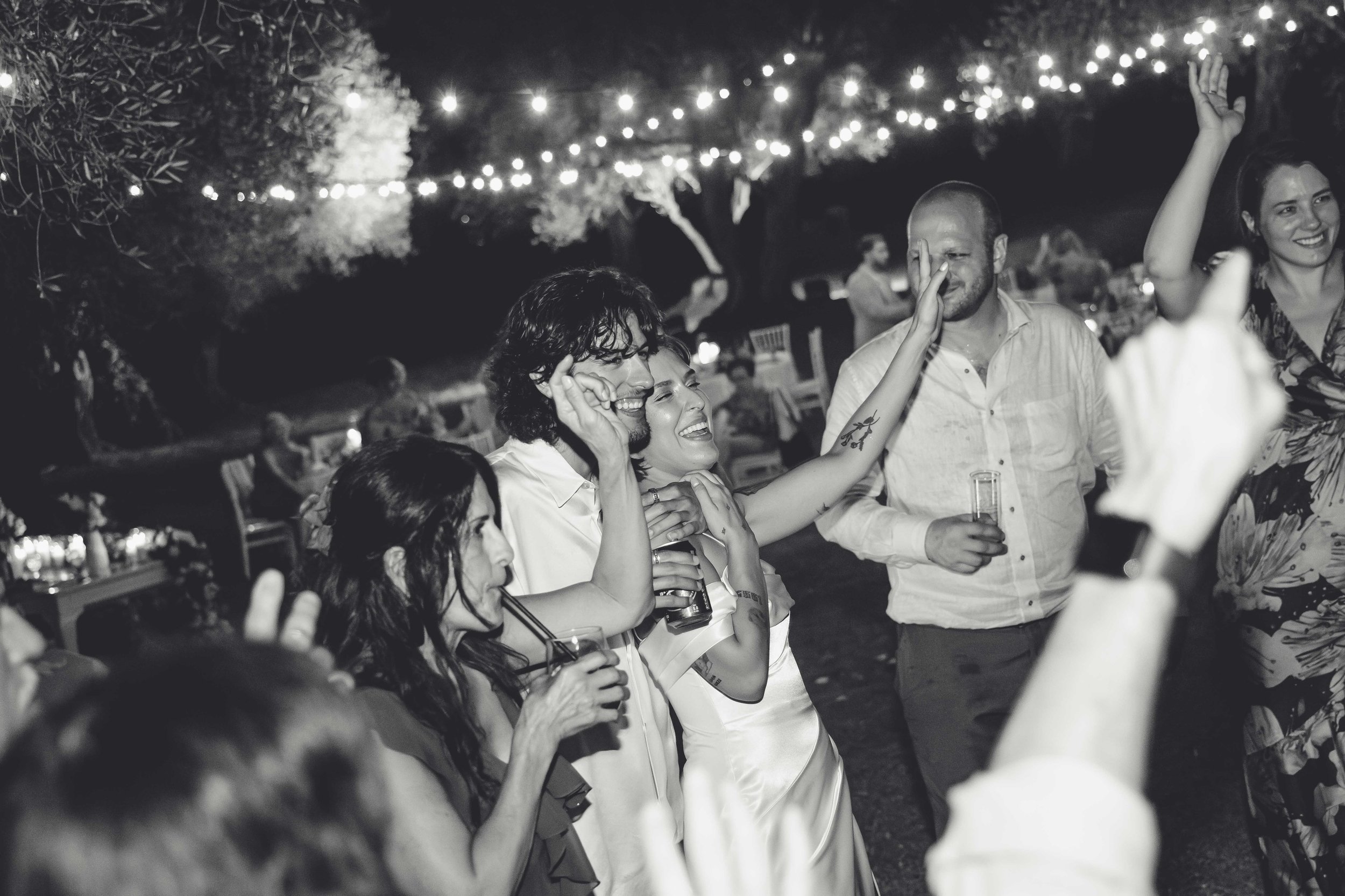 People celebrating at a nighttime outdoor event with string lights overhead. Several people are smiling and holding drinks, engaging in joyful conversation and dancing.