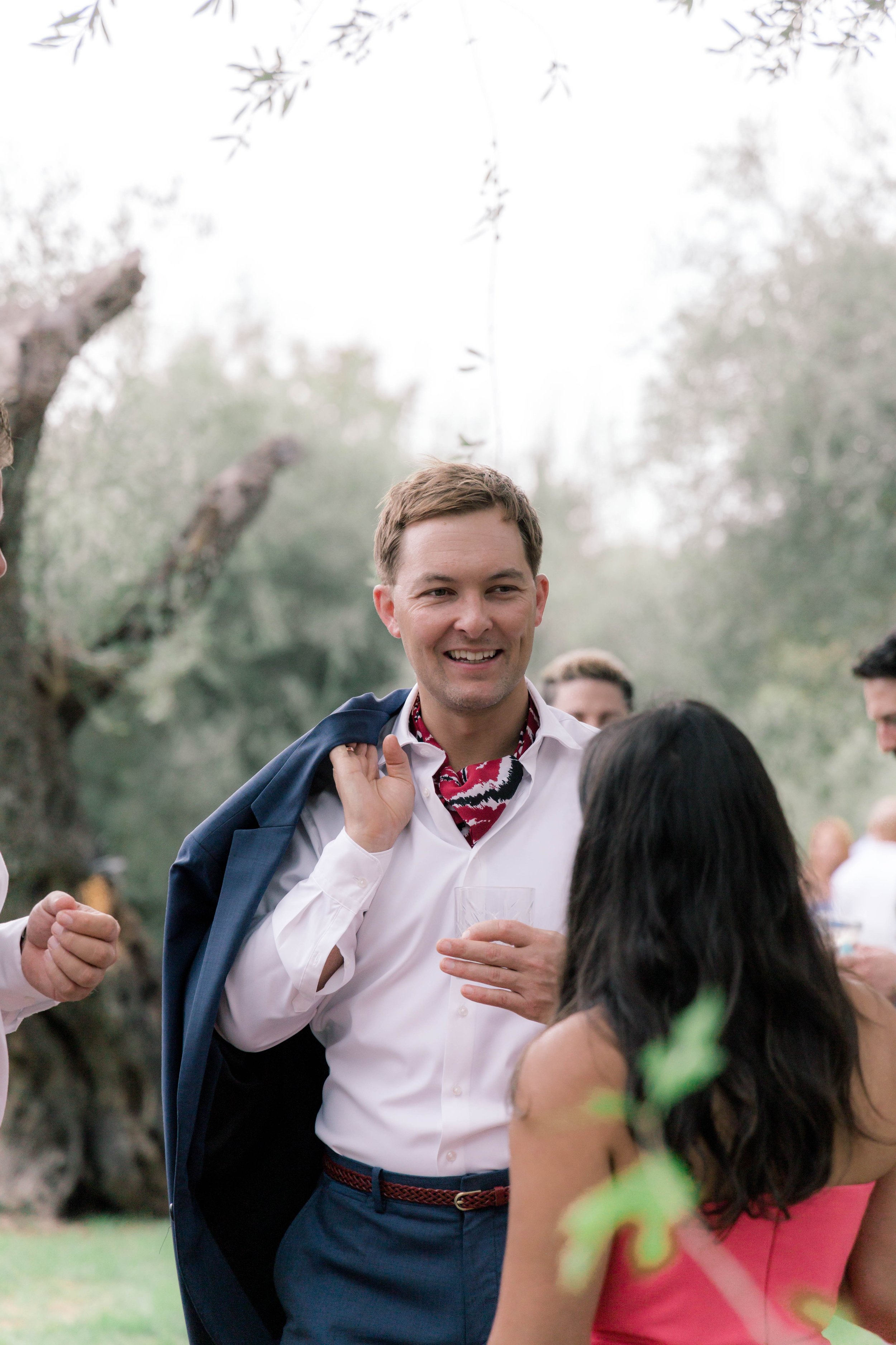 A man in a white shirt and carrying a dark jacket over his shoulder, smiling and talking to a woman at an outdoor social gathering with trees in the background.