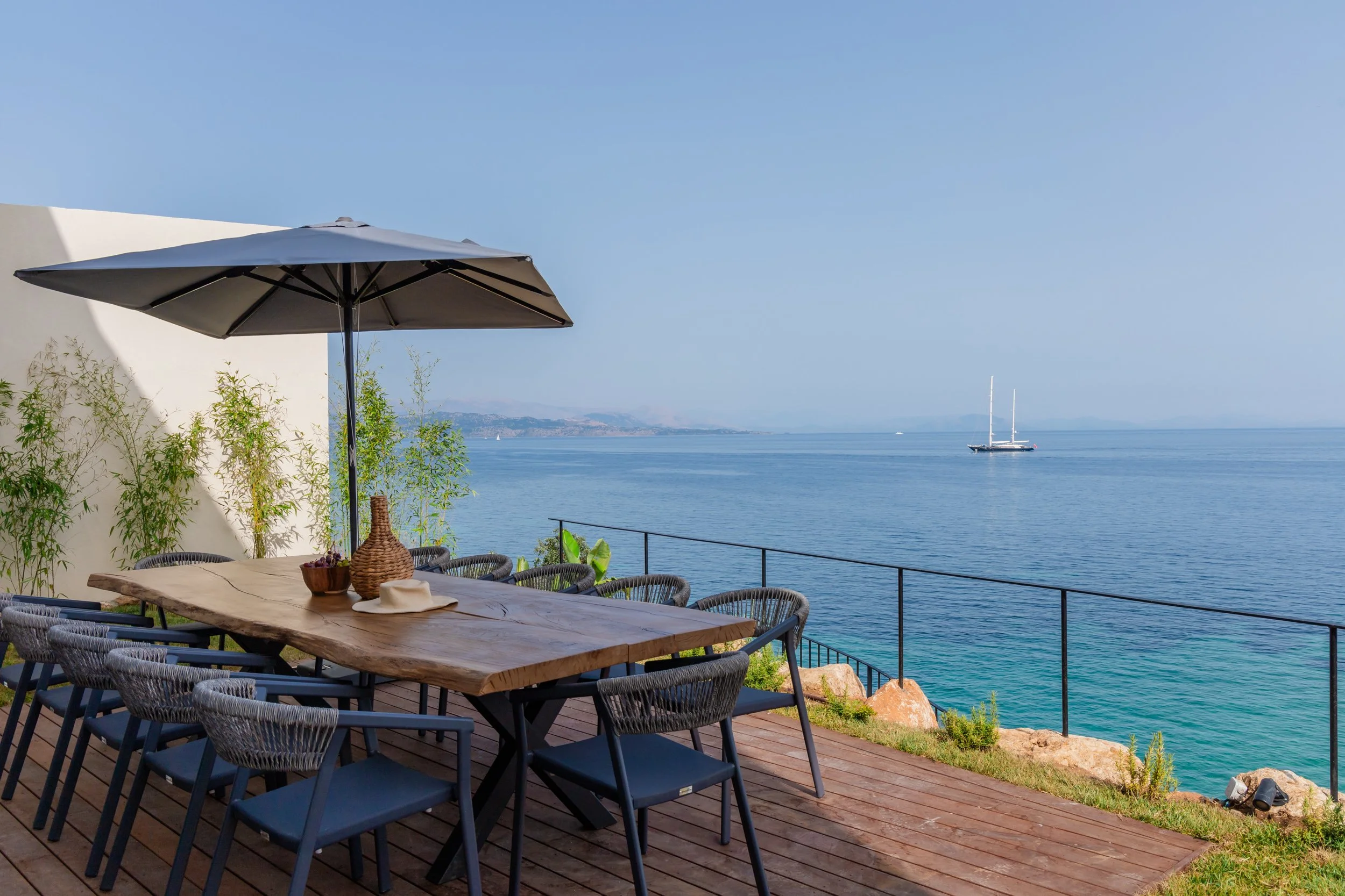 Outdoor dining area on wooden deck overlooking calm ocean with sailboat in distance, white wall with greenery, and a large patio umbrella.
