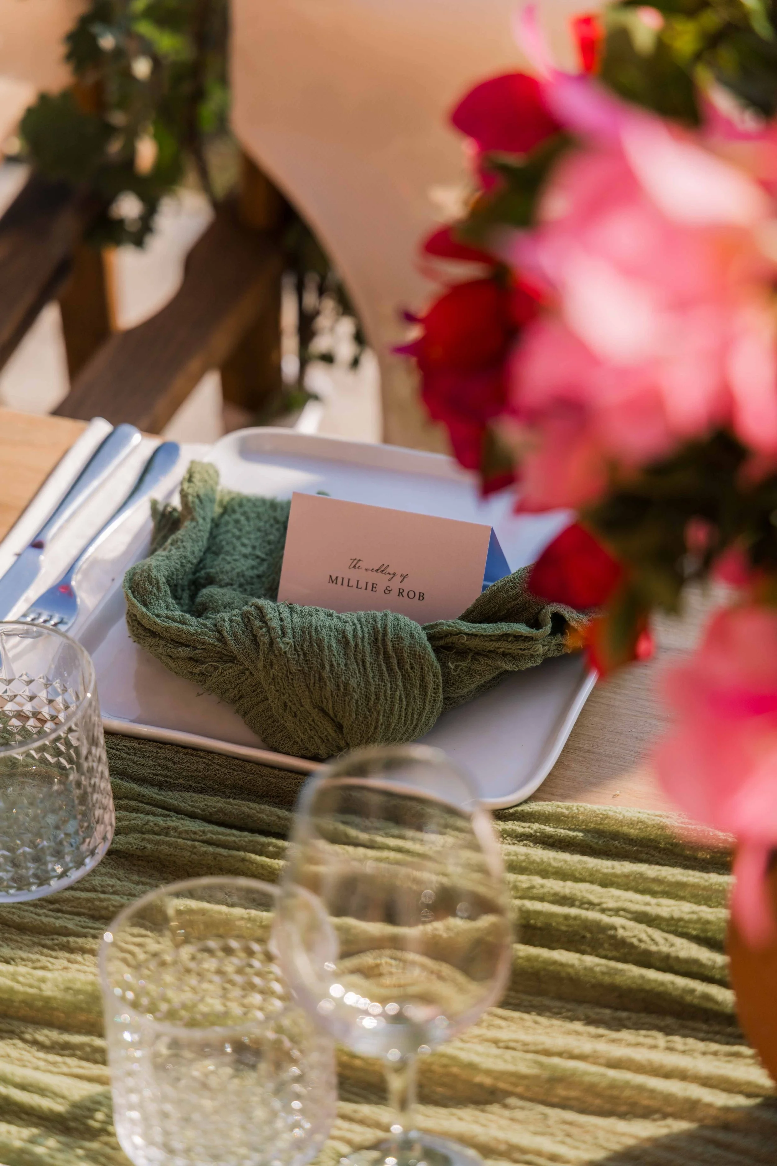 A decorated table setting for a wedding, featuring a white plate with a green cloth napkin and a personalized place card reading 'the wedding of MILLIE & ROB,' surrounded by glassware, a cutlery set, and flowers in the foreground.