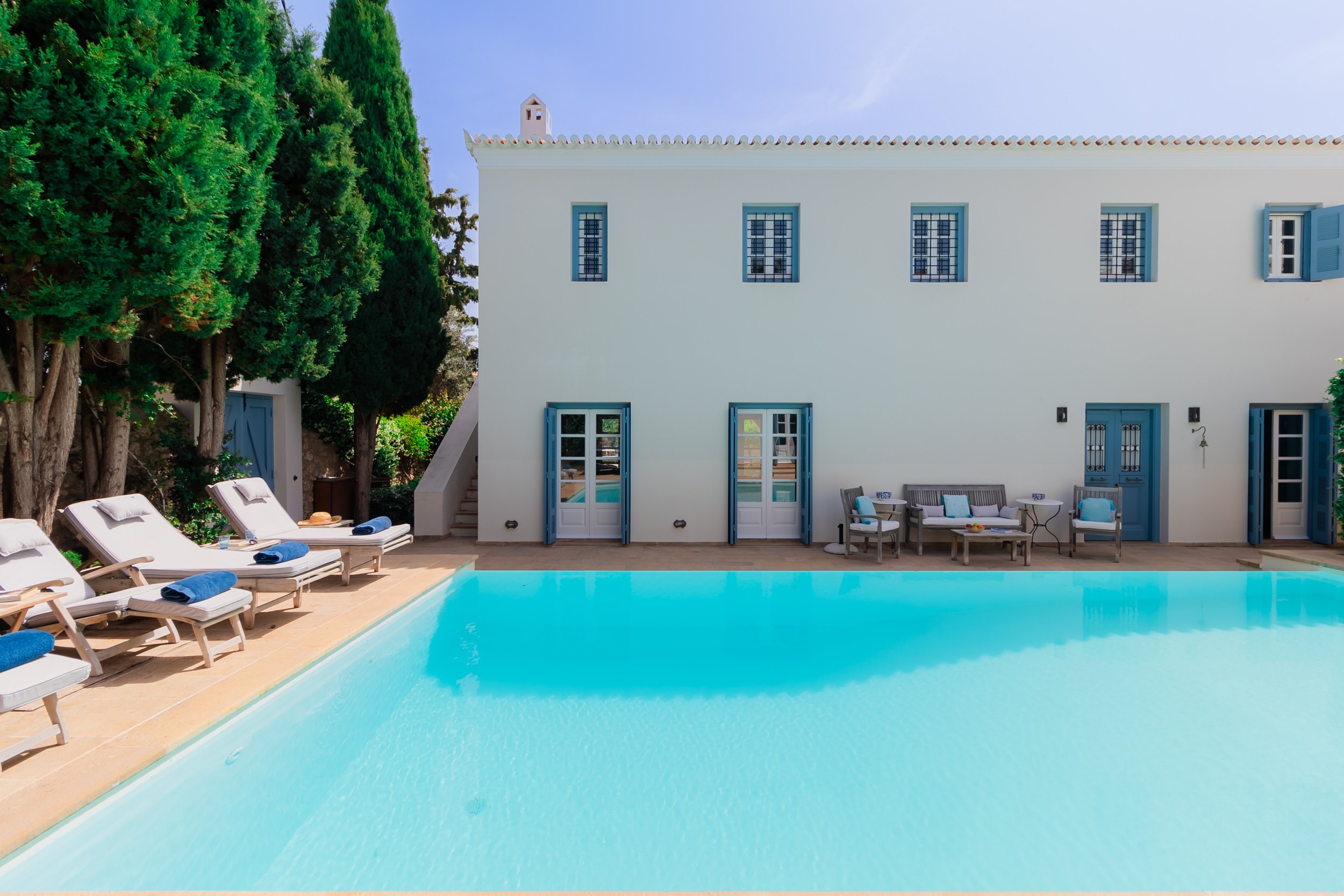 A white Mediterranean-style house with blue shutters and doors, overlooking a swimming pool with lounge chairs and outdoor seating, surrounded by tall green trees.