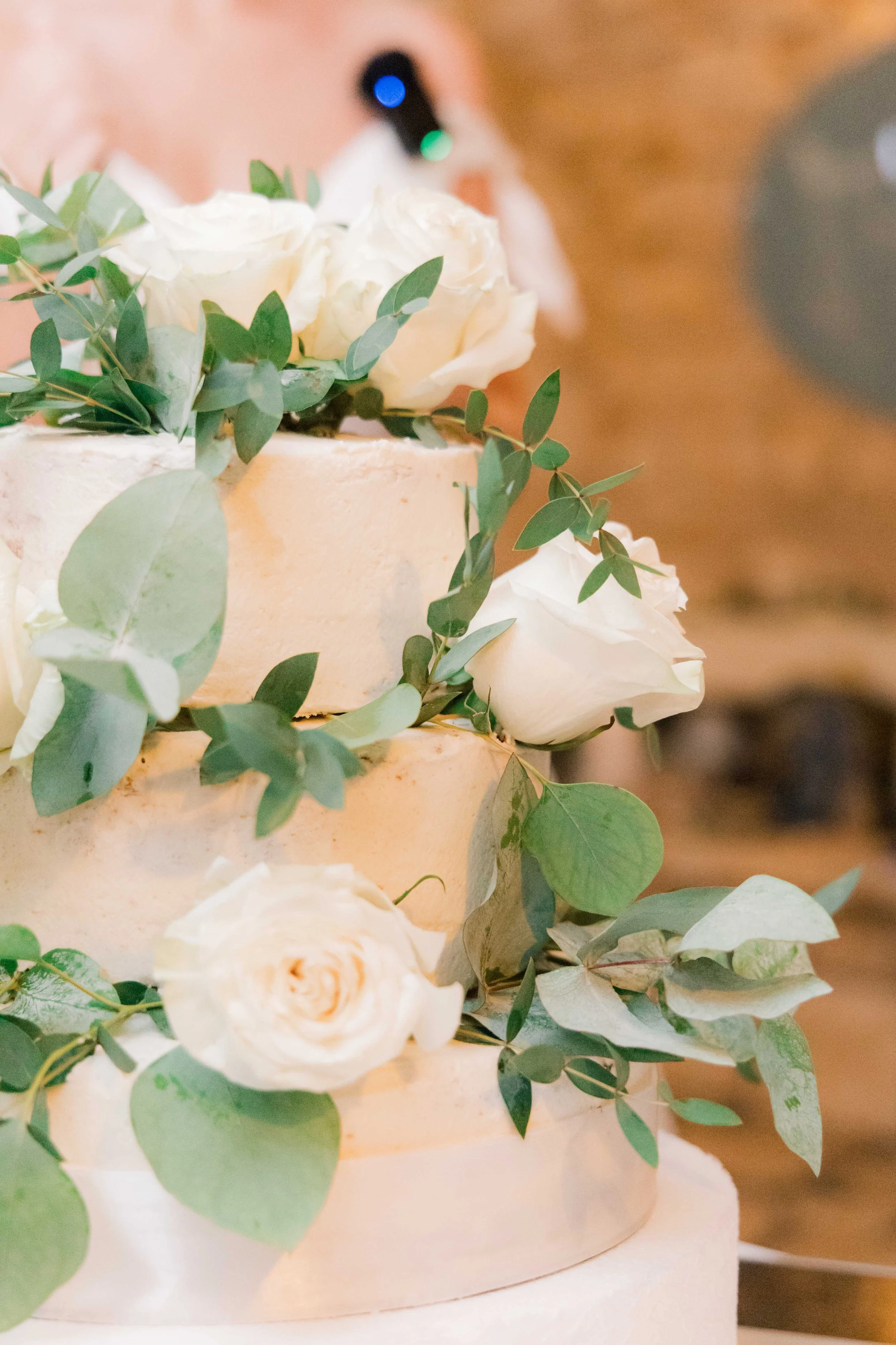 Close-up of a three-tiered wedding cake decorated with white roses and green eucalyptus leaves.