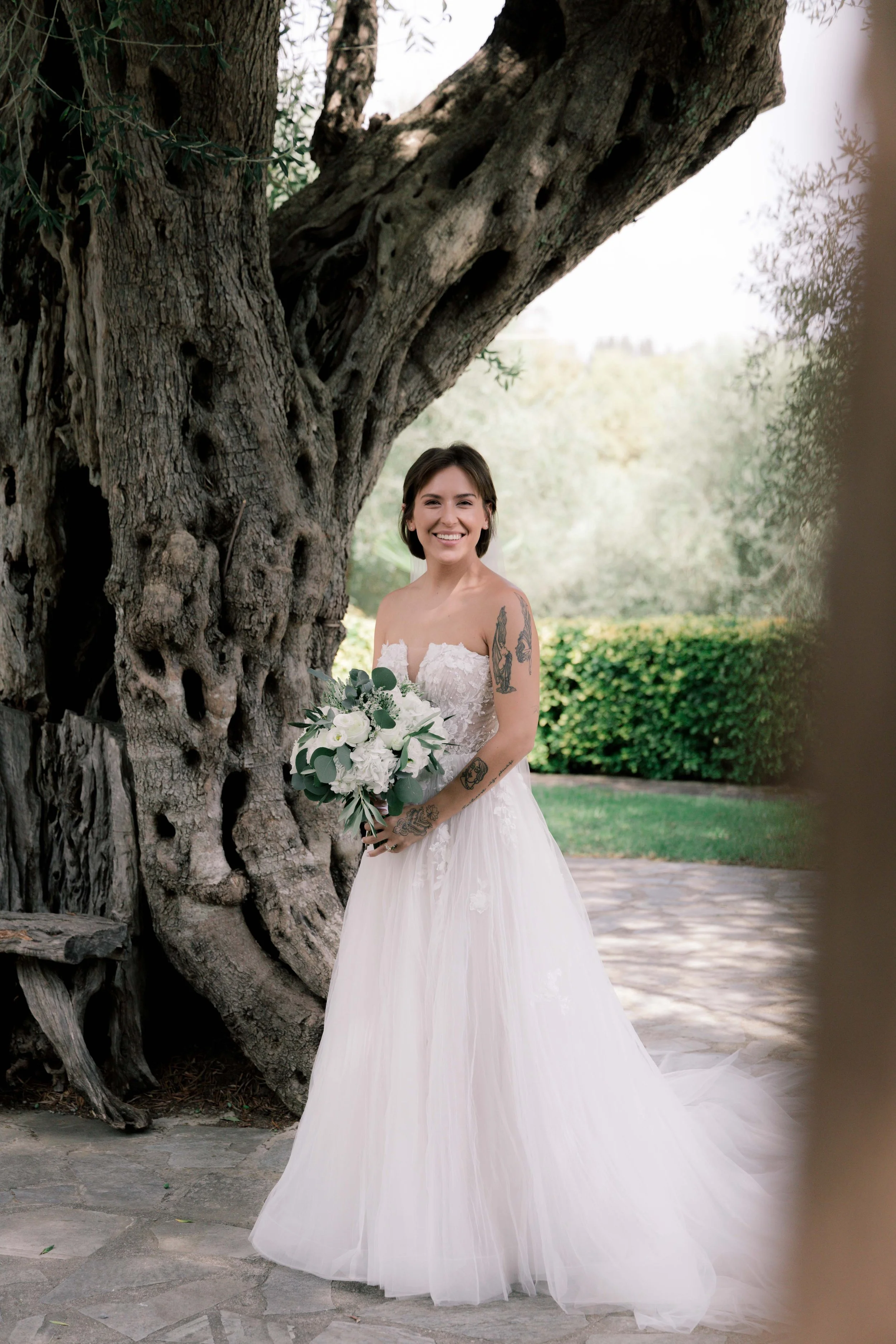 A smiling bride with short brown hair in a strapless white wedding gown holding a bouquet of white flowers, standing outdoors near a large, old tree with textured bark, in a garden setting.