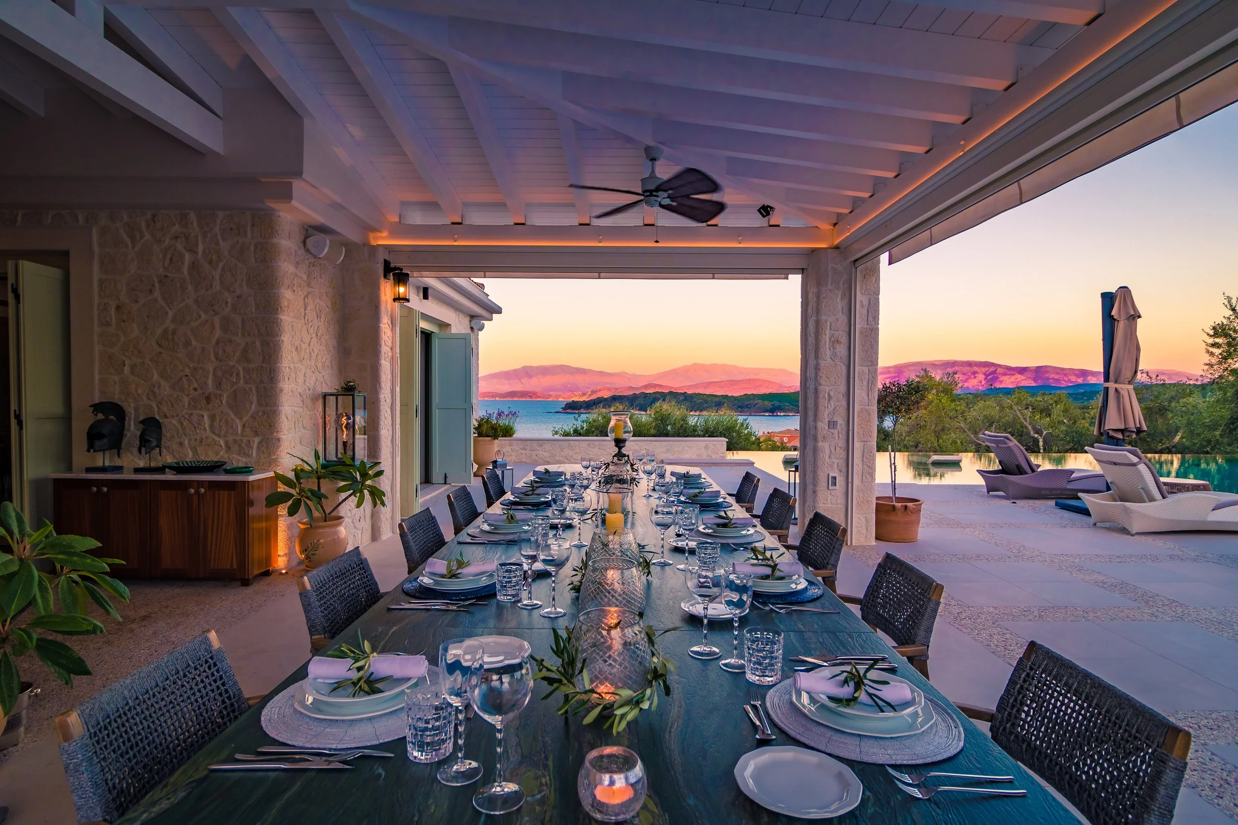 Outdoor patio dining area with a long table set for a meal, overlooking a body of water with mountains in the distance during sunset.