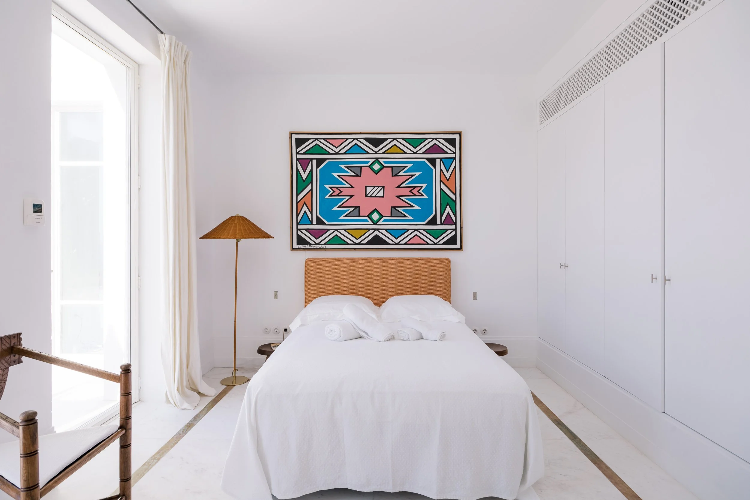 A minimalist bedroom with white walls and a bed with a wicker headboard, white bedding, and rolled white towels. There's a colorful geometric painting above the bed, a wooden bedside table on each side, a tall floor lamp with a woven shade, and a lar