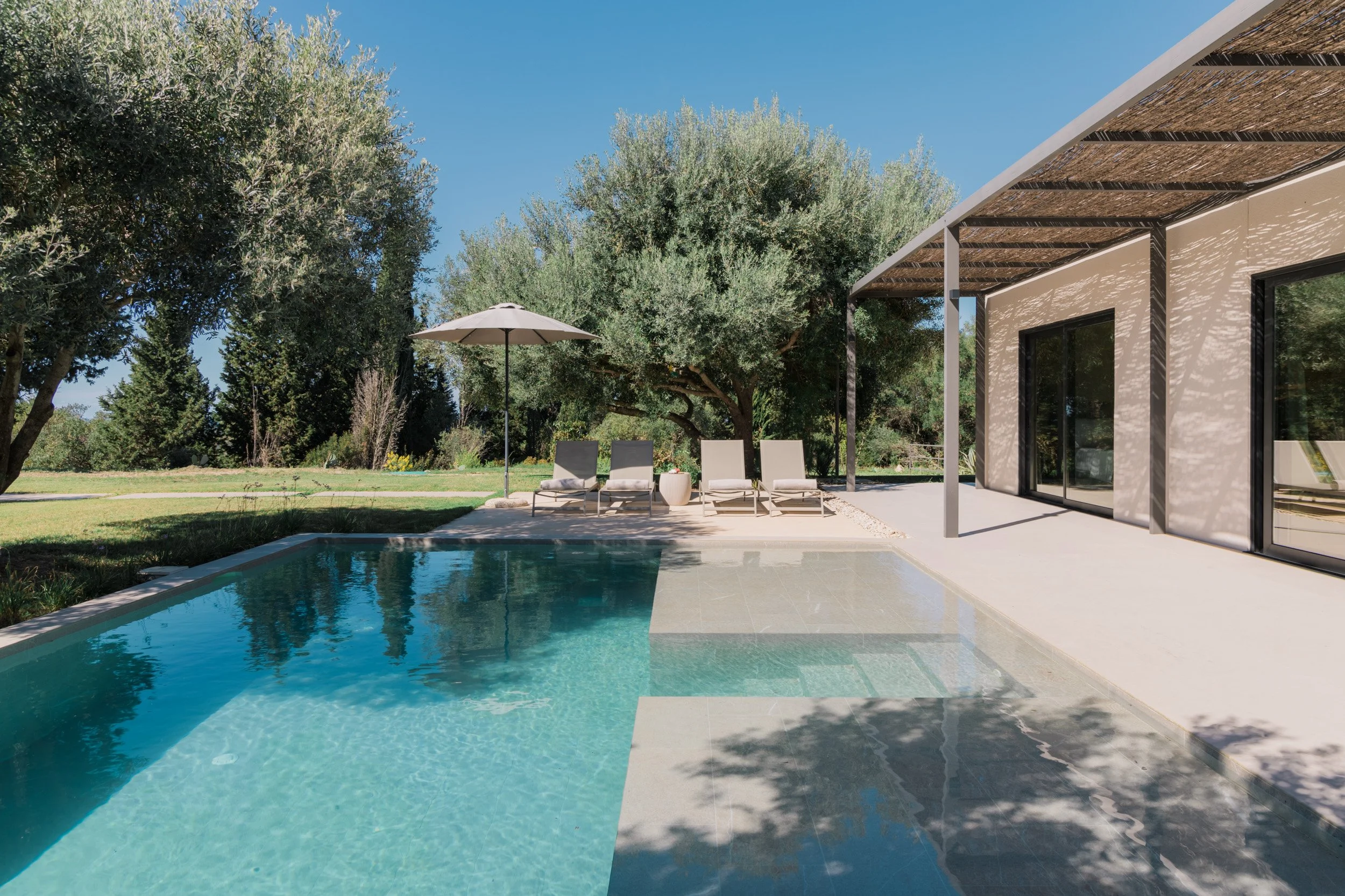 Outdoor scene featuring a swimming pool with clear blue water, surrounded by lawn, trees, and garden chairs. There is a patio with a modern house that has large sliding glass doors and a wooden pergola.