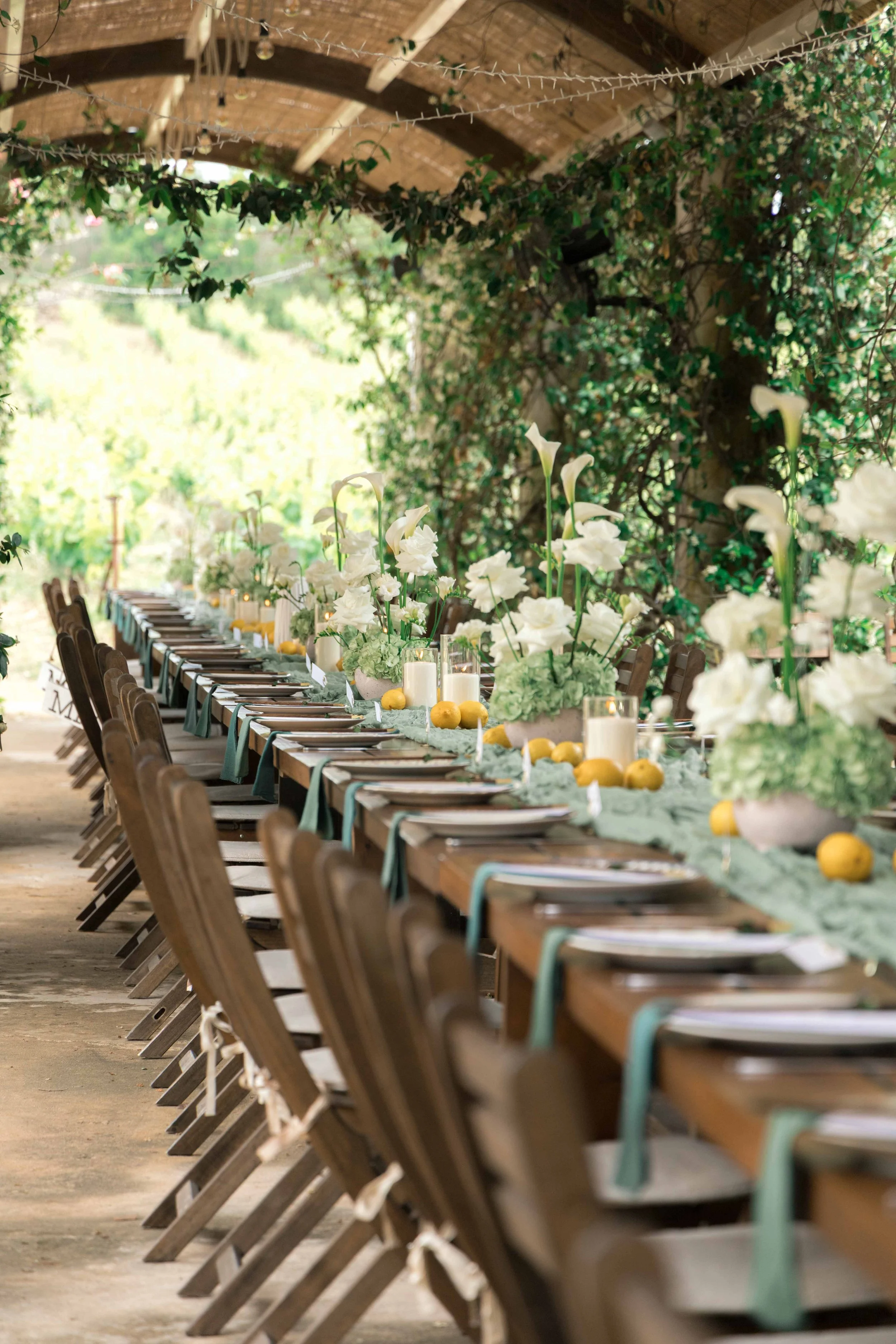 Long wooden table decorated with white flowers, greenery, candles, and lemons, set up for an outdoor event under a roof with hanging string lights and abundant green foliage.