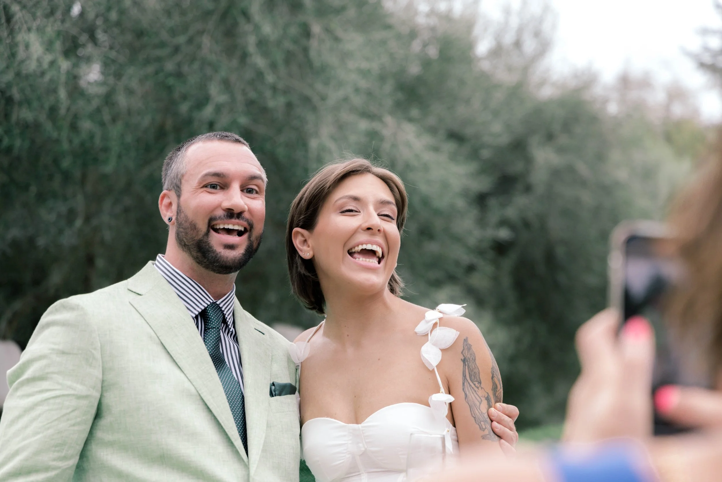 A smiling man and woman taking a photo together outdoors, woman has a tattoo on her arm and is wearing a white dress, man is wearing a beige suit with a checkered shirt and tie, background features trees.