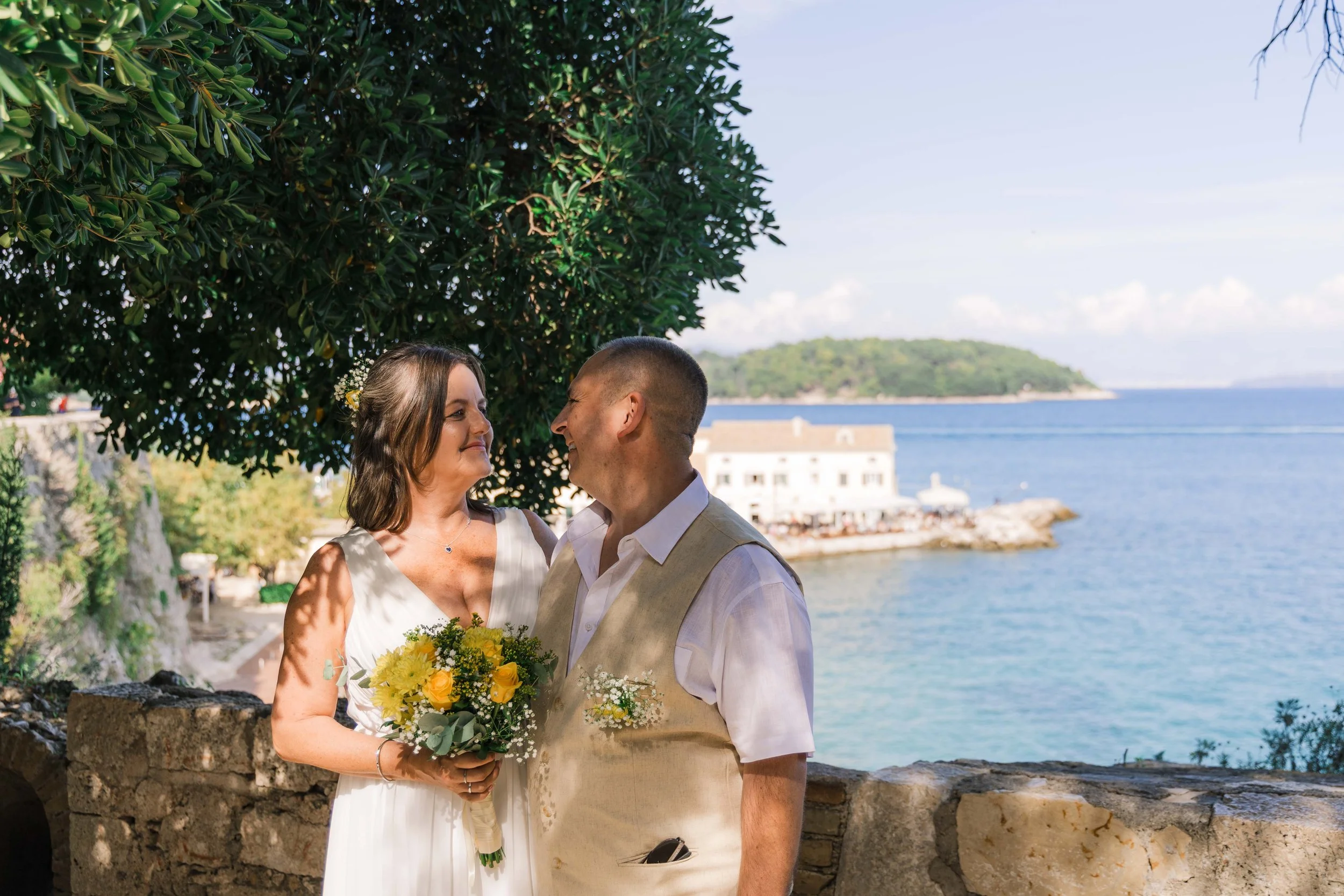 A bride and groom look at each other smiling, standing outdoors near a stone wall with trees and water in the background, during a wedding celebration.